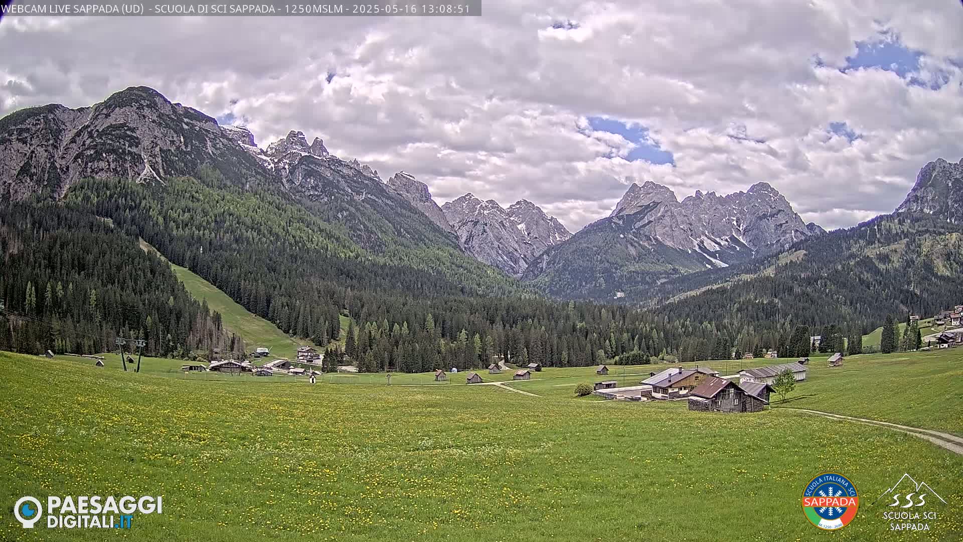 A partly cloudy day reveals a valley of green meadows and low-lying buildings nestled beneath a mountain range with patches of snow.