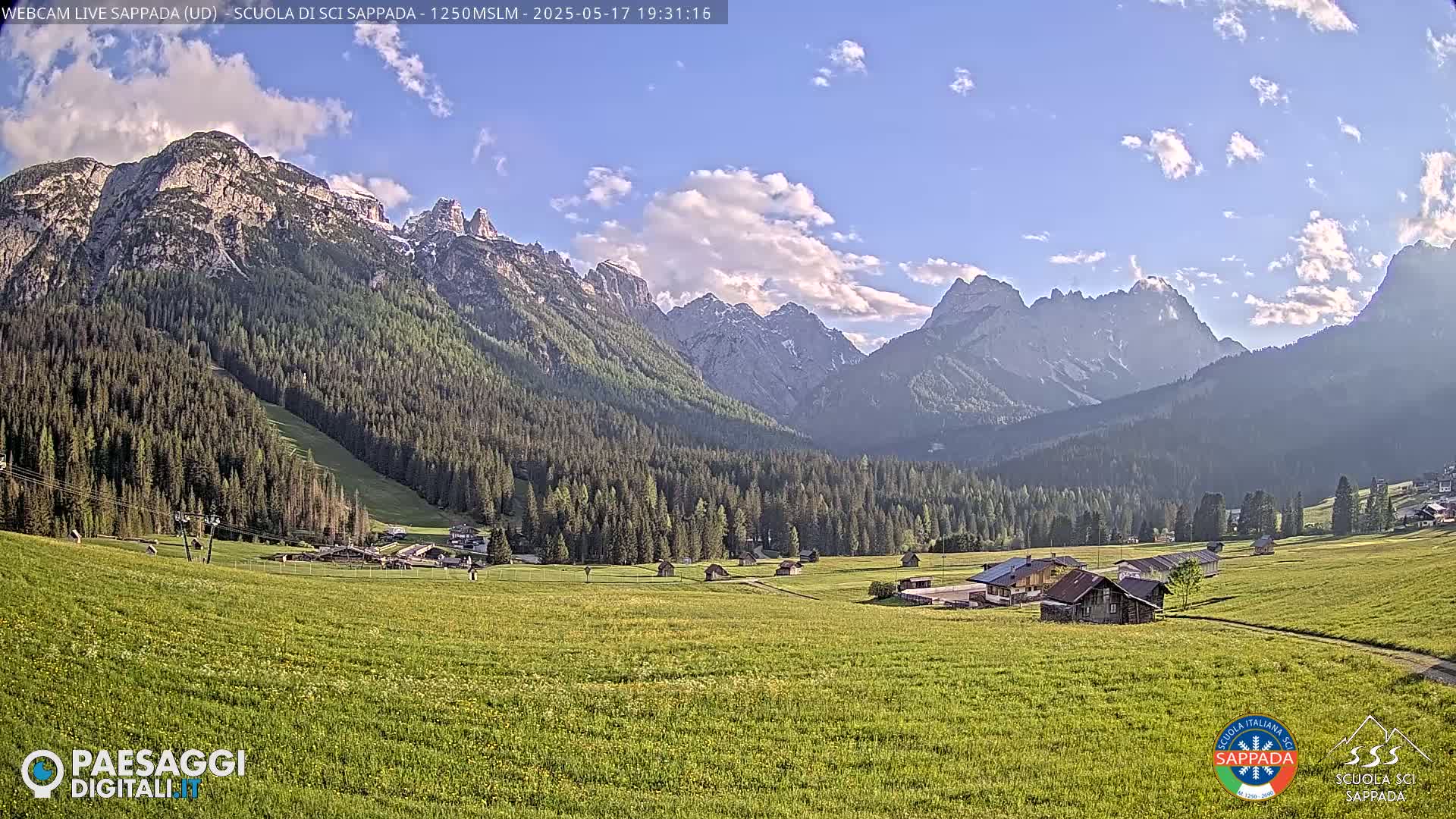 A sunny valley with green fields and a few buildings is nestled between towering, snow-capped mountains under a partly cloudy sky.