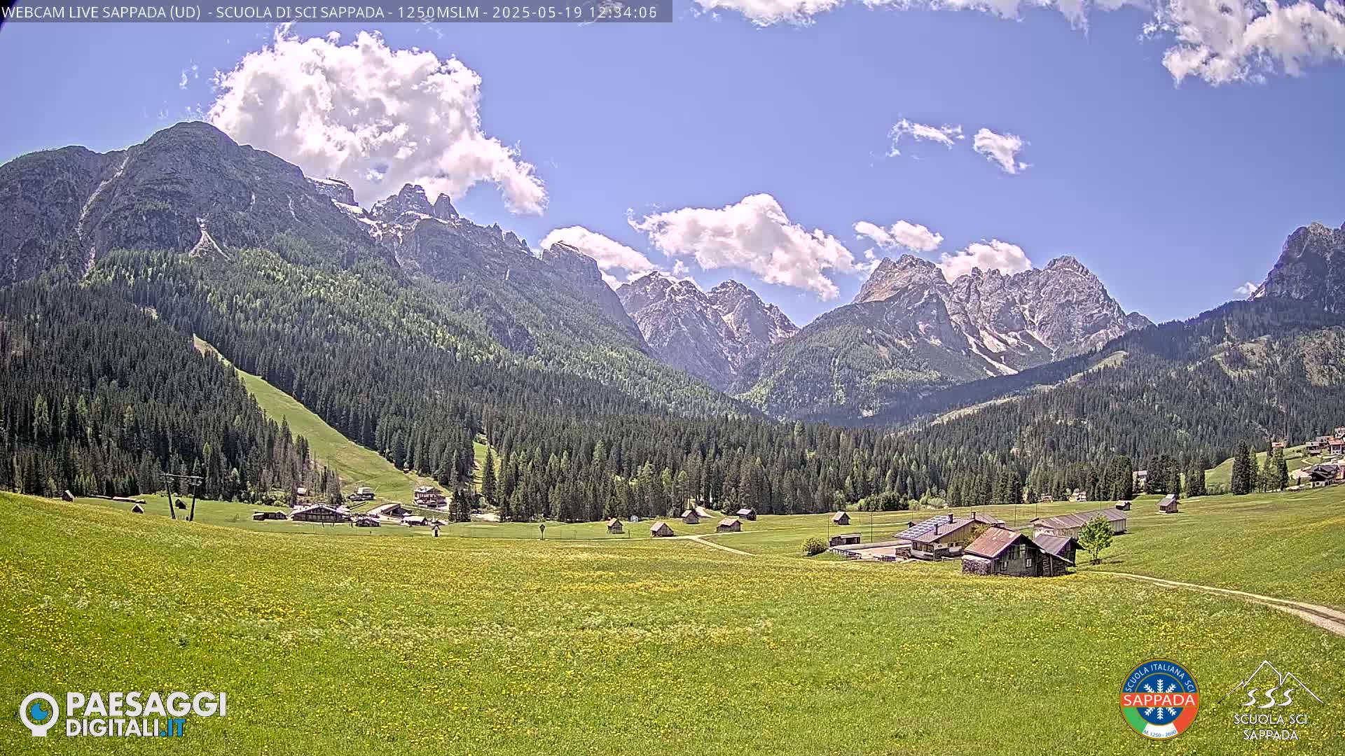 A sunny day reveals a valley of green fields dotted with small buildings, nestled beneath a towering range of snow-capped mountains.