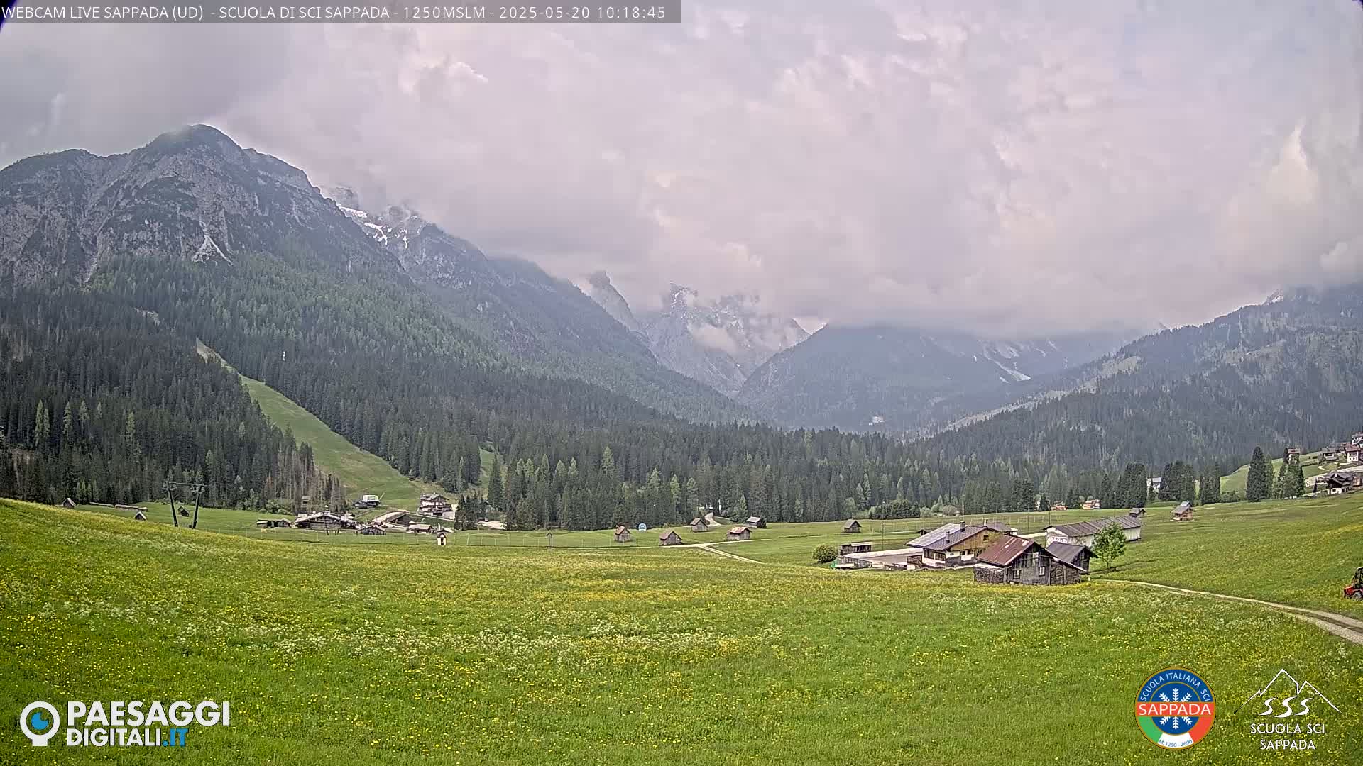 A valley dotted with small buildings and green fields is nestled beneath a cloudy sky and mountains partially covered in snow.