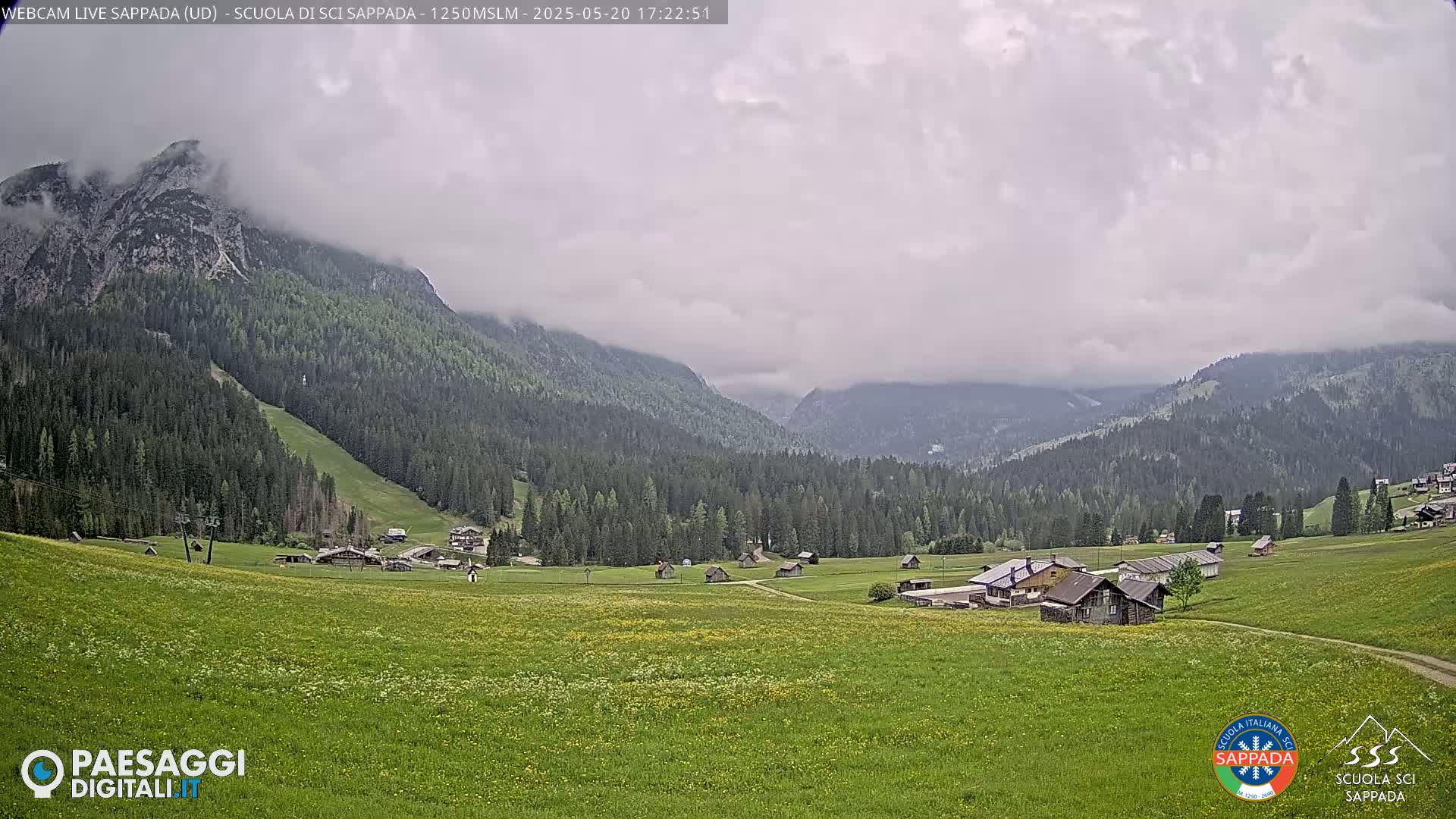 A valley dotted with small buildings and lush green fields is nestled between mountains partially shrouded in clouds under an overcast sky.