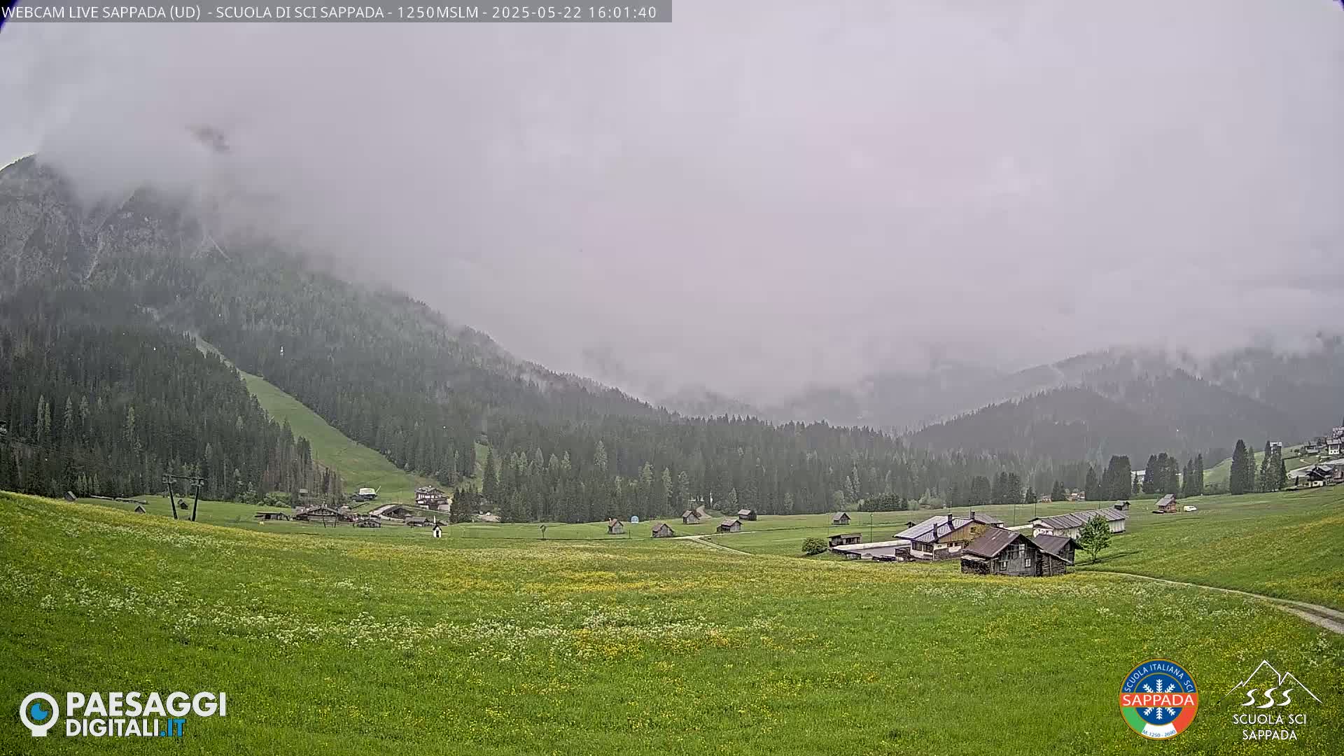 A misty, overcast day reveals a valley dotted with small buildings and green fields, backed by a mountain range partially shrouded in fog.