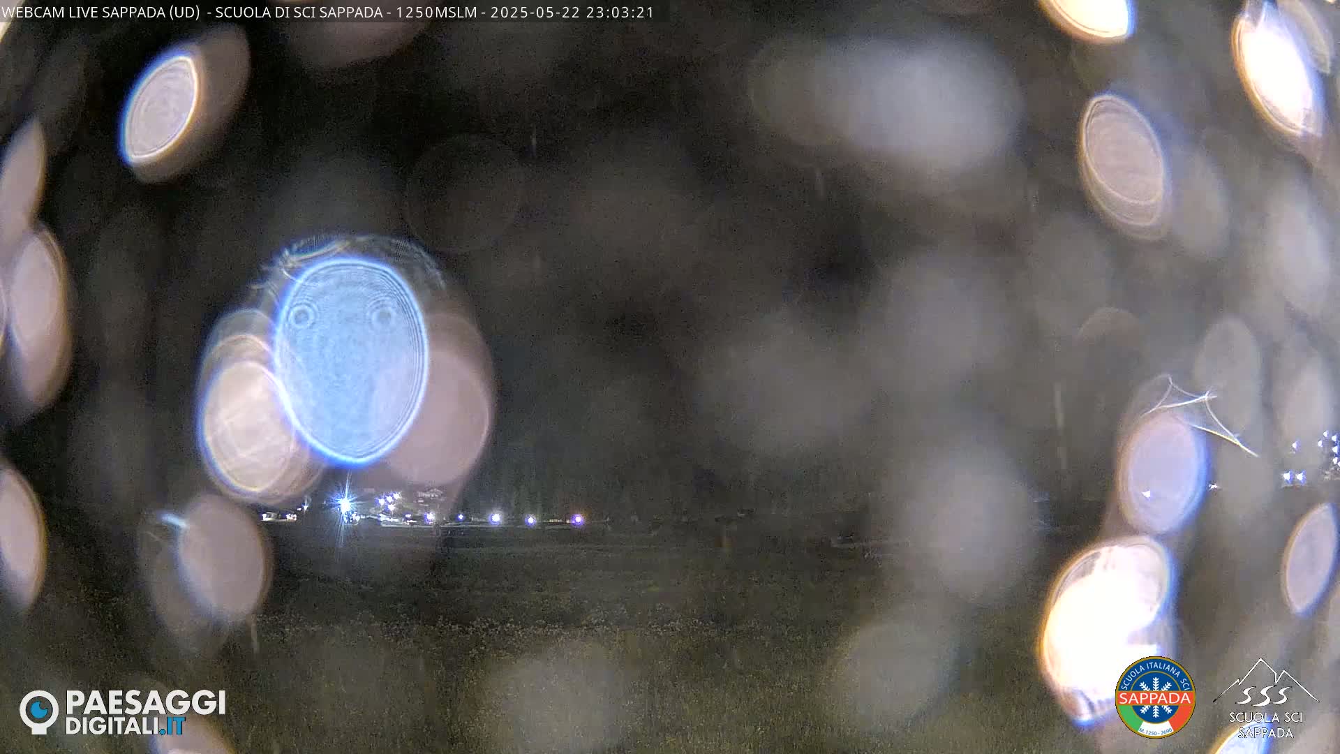 A blurry nighttime image shows out-of-focus lights in the distance, obscured by water droplets on a lens.