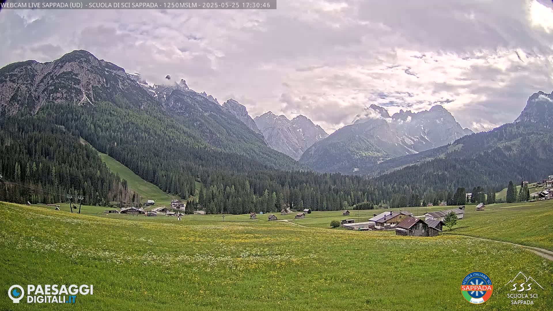 A valley dotted with small buildings and wildflowers stretches out beneath a partly cloudy sky, framed by a mountain range with some snow cover.