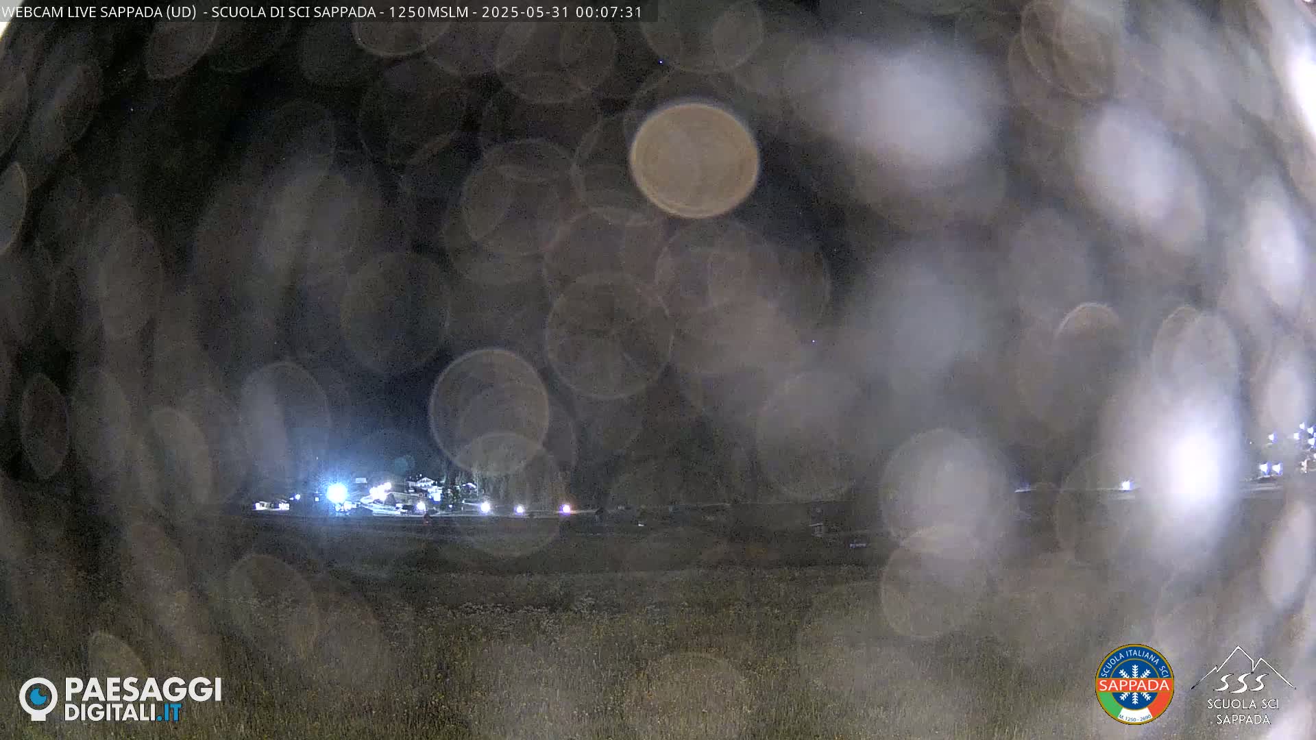 A nighttime view of a dimly lit area, possibly a parking lot or roadway, obscured by blurry raindrops or condensation on the camera lens.