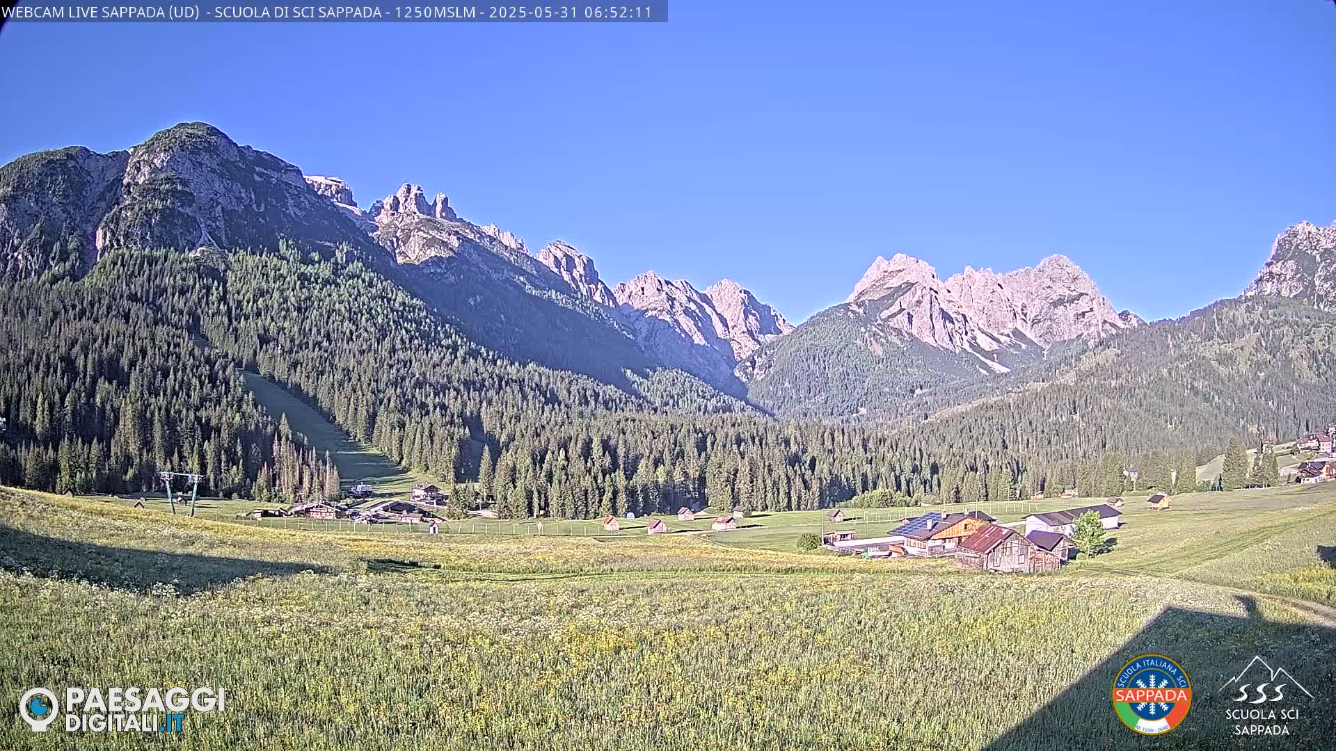 A mountain valley with a grassy field in the foreground, a small village nestled in the mid-ground, and a large mountain range in the background under a clear blue sky.