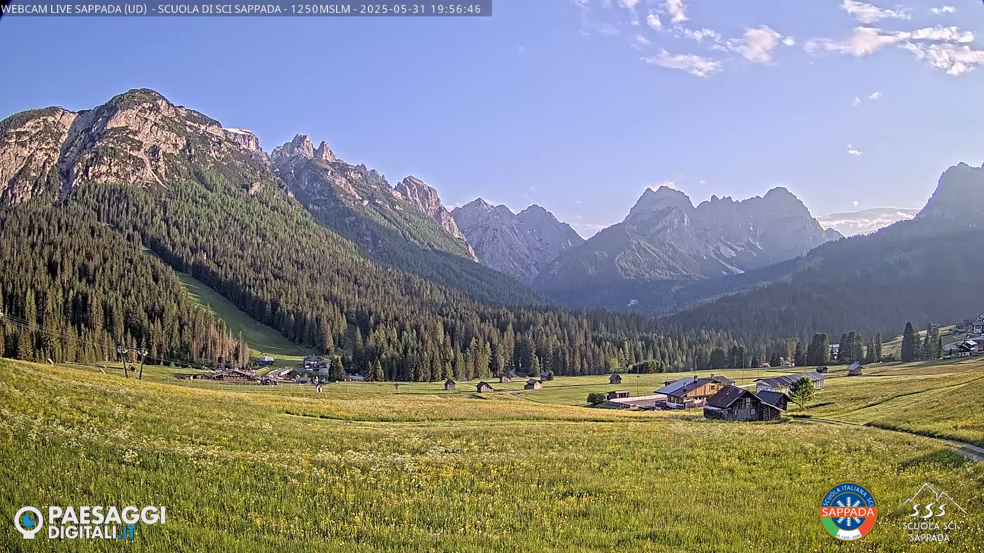 A valley filled with wildflowers and a few buildings is nestled between large, rocky mountains under a mostly clear, bright sky.