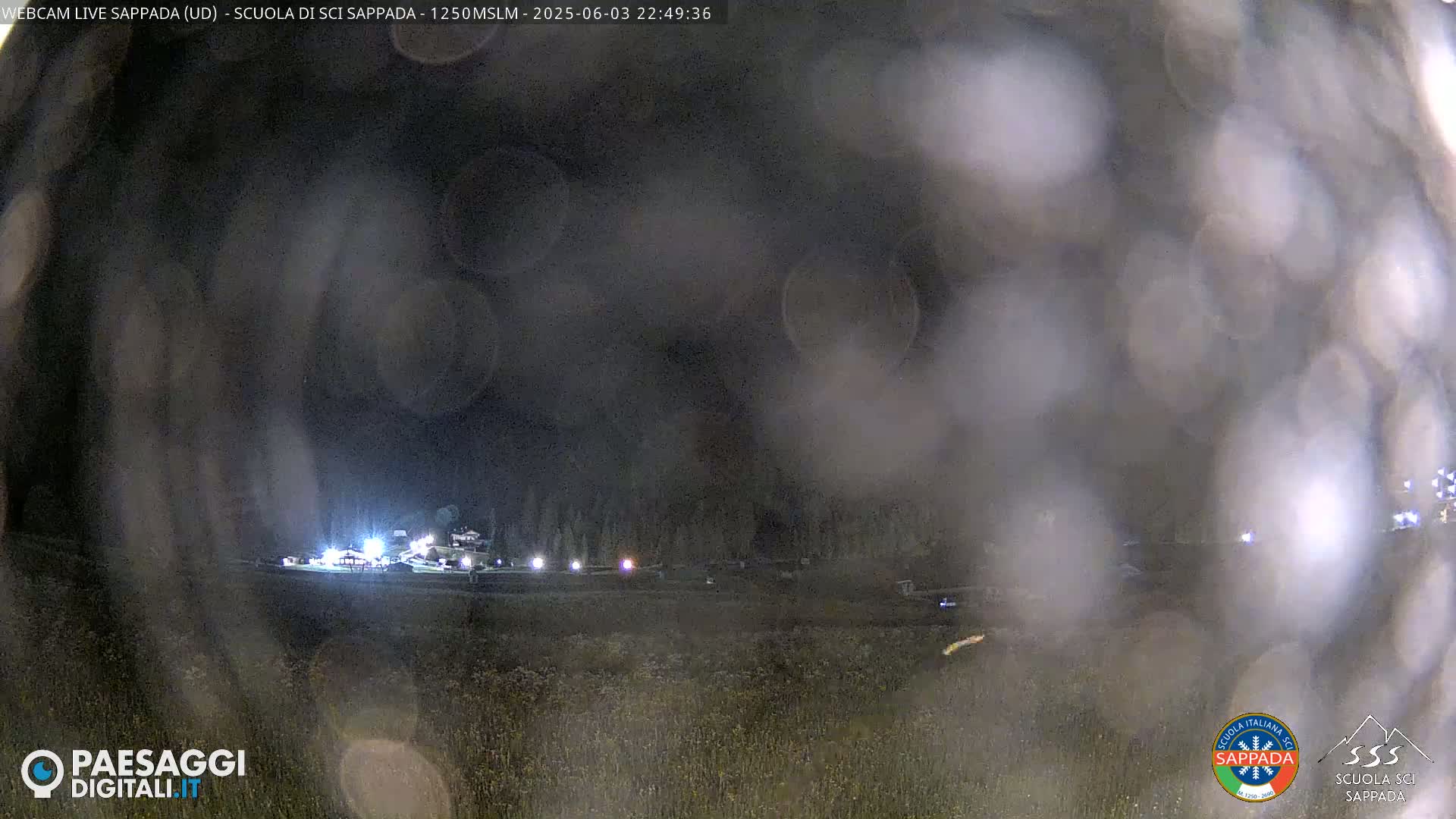 A nighttime, blurry view of a dimly lit area with some structures and vegetation, possibly under light rain or fog.
