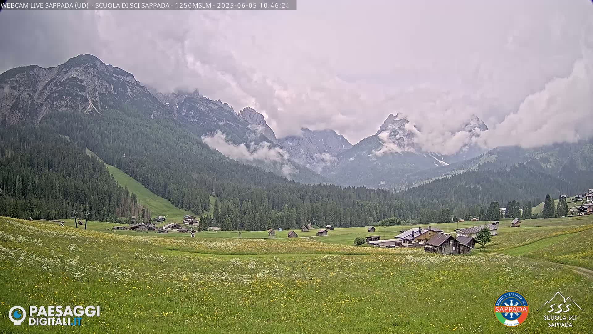 A picturesque valley dotted with small buildings and wildflowers stretches out beneath a cloudy sky, framed by imposing, cloud-covered mountains.