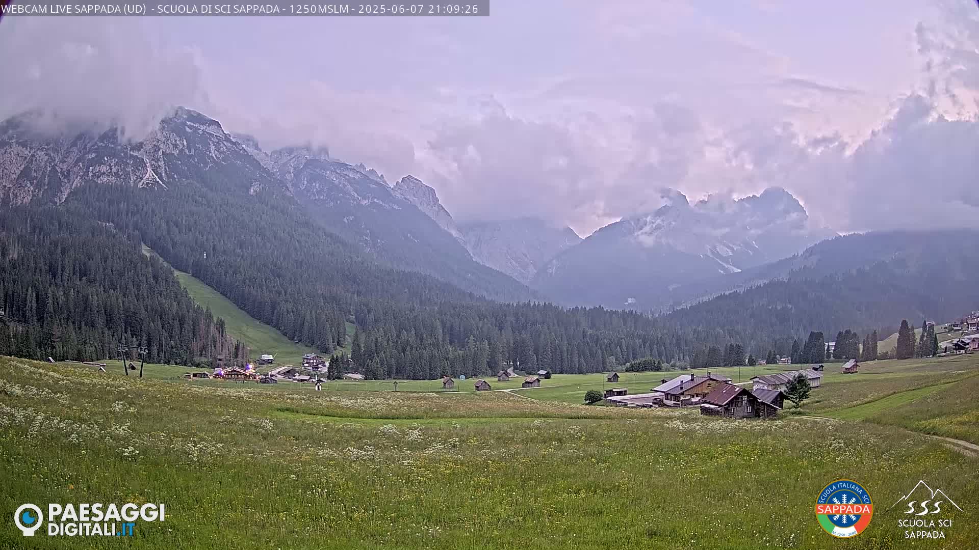 A mountain valley with a few small buildings and a field of wildflowers is partly obscured by clouds under an overcast sky.