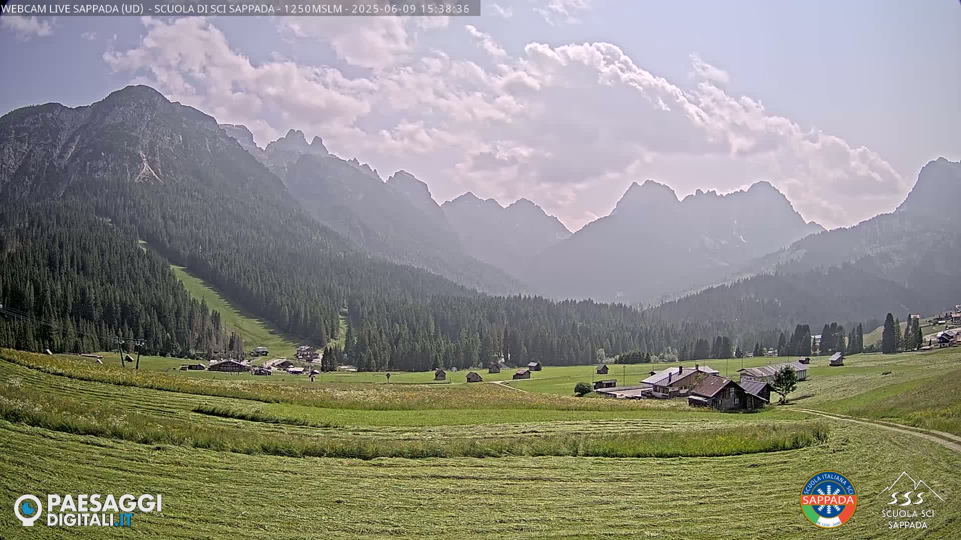 A partly cloudy day reveals a valley with freshly cut hay fields, a few small buildings, and a dense evergreen forest leading up to a hazy mountain range.
