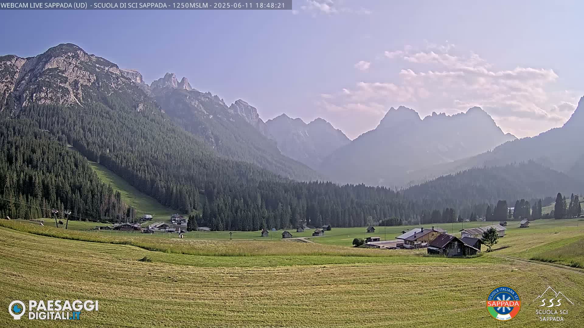 A hazy, sunny day reveals a valley village nestled among verdant fields and dark green forests, with a mountain range in the background.