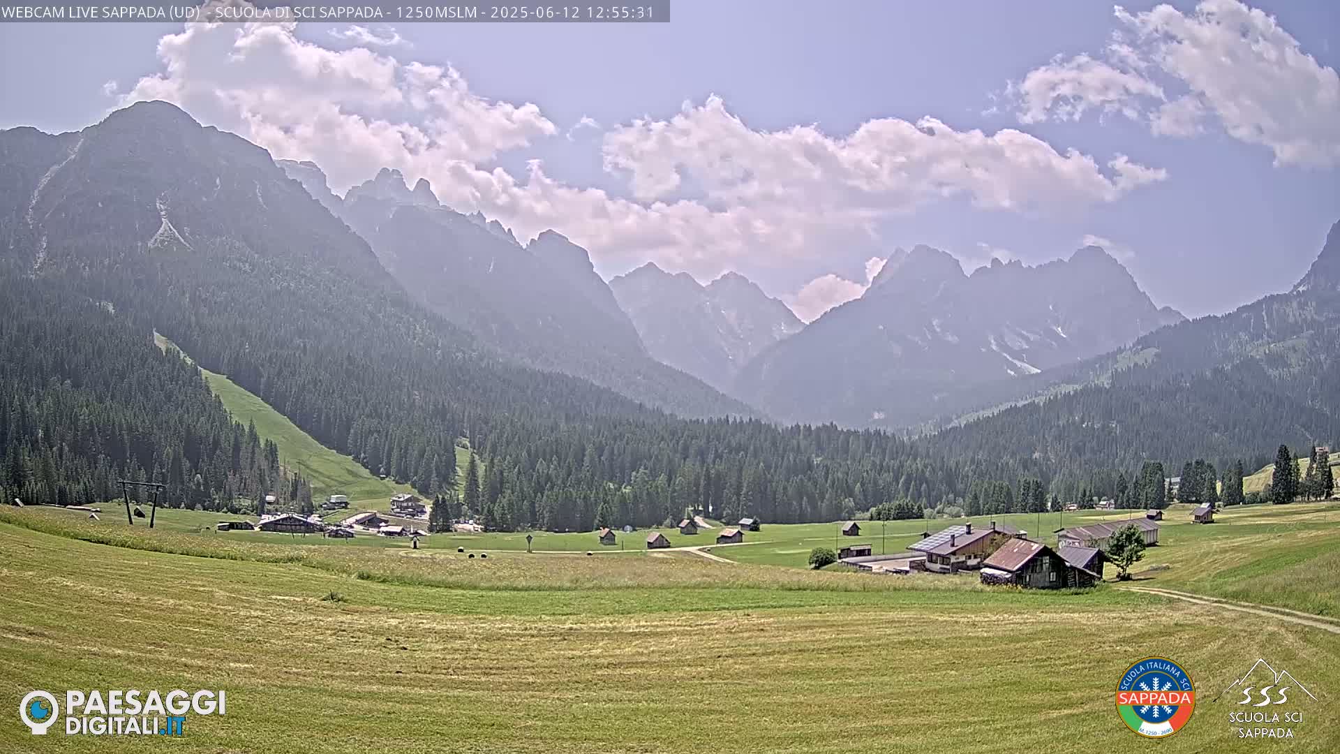 A sunny day in a mountain valley shows a small village nestled among green fields and evergreen forests, with a range of rugged, hazy mountains in the background.