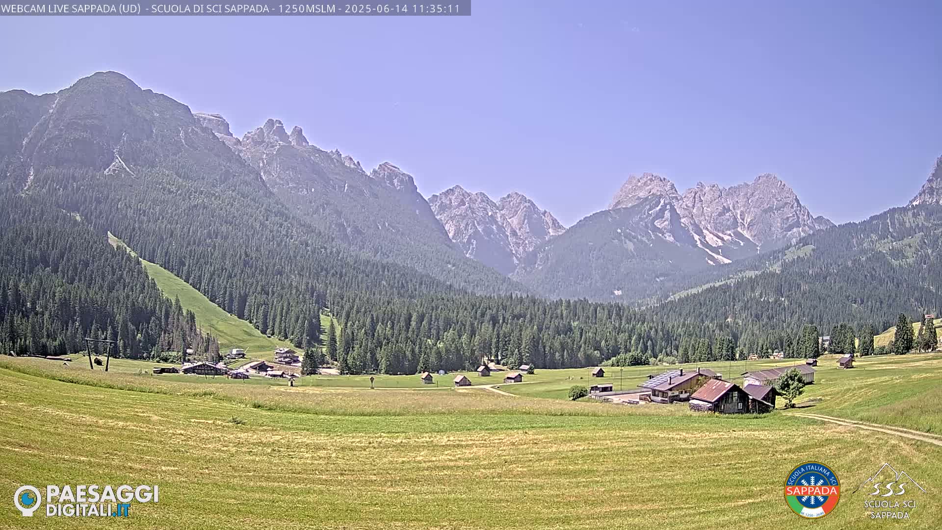 A sunny day reveals a valley of green fields and small buildings nestled among a dark forest, all overlooked by a majestic mountain range under a clear sky.