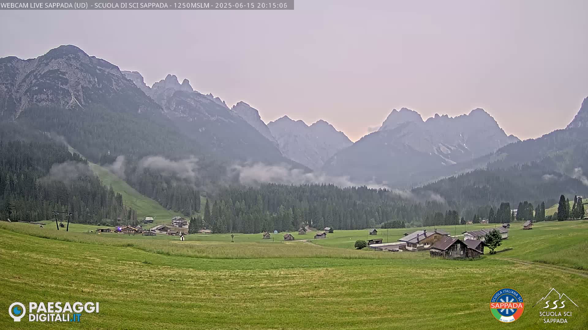 A valley with low-lying fog partially obscuring a verdant field dotted with buildings, set against a backdrop of misty mountains under a pale, dusky sky.