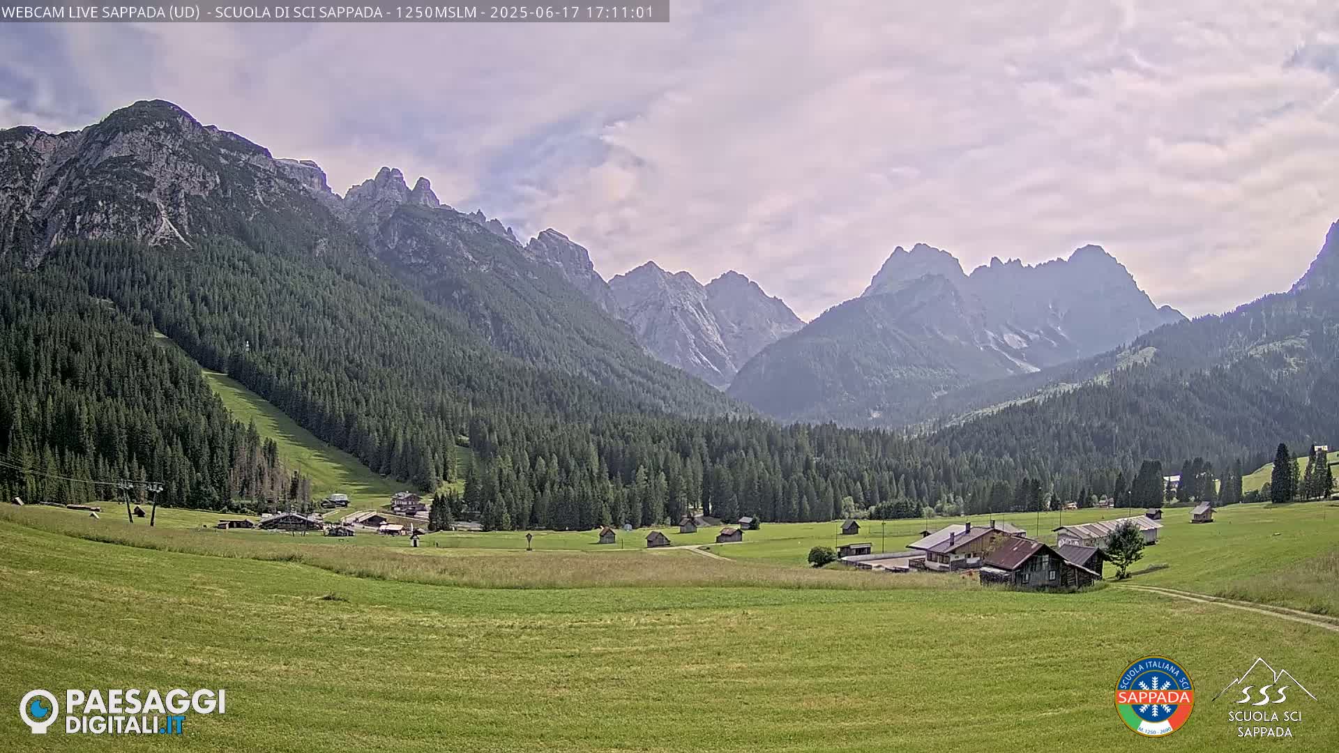 A valley with green fields and a small village is nestled at the base of a mountain range under a mostly cloudy sky.