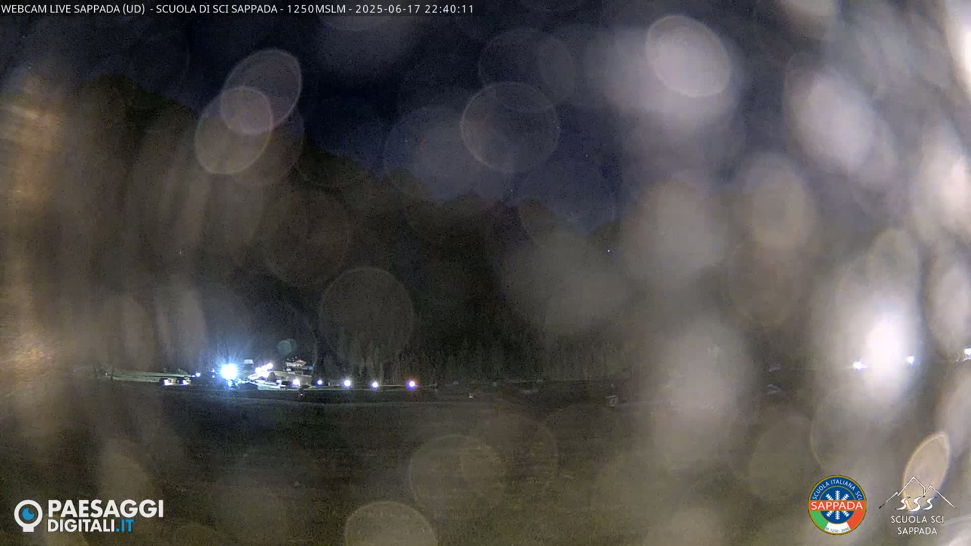 A nighttime view of a dimly lit area, possibly a parking lot or roadway, with a dark mountain range in the background, seen through a rain-streaked or fogged surface.