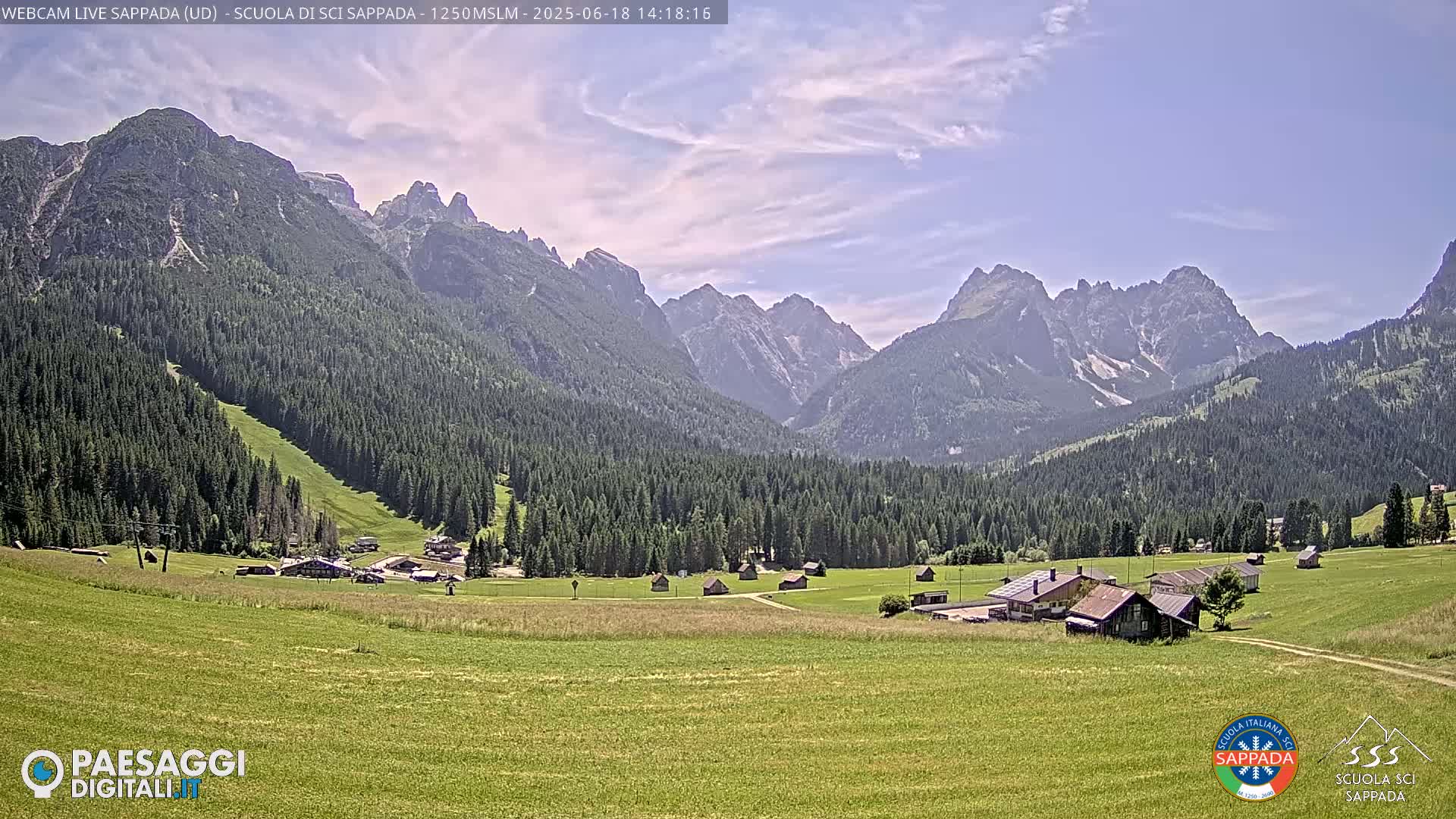 A sunny day in a mountainous valley shows a green meadow with small buildings, a coniferous forest, and a range of jagged peaks in the background.