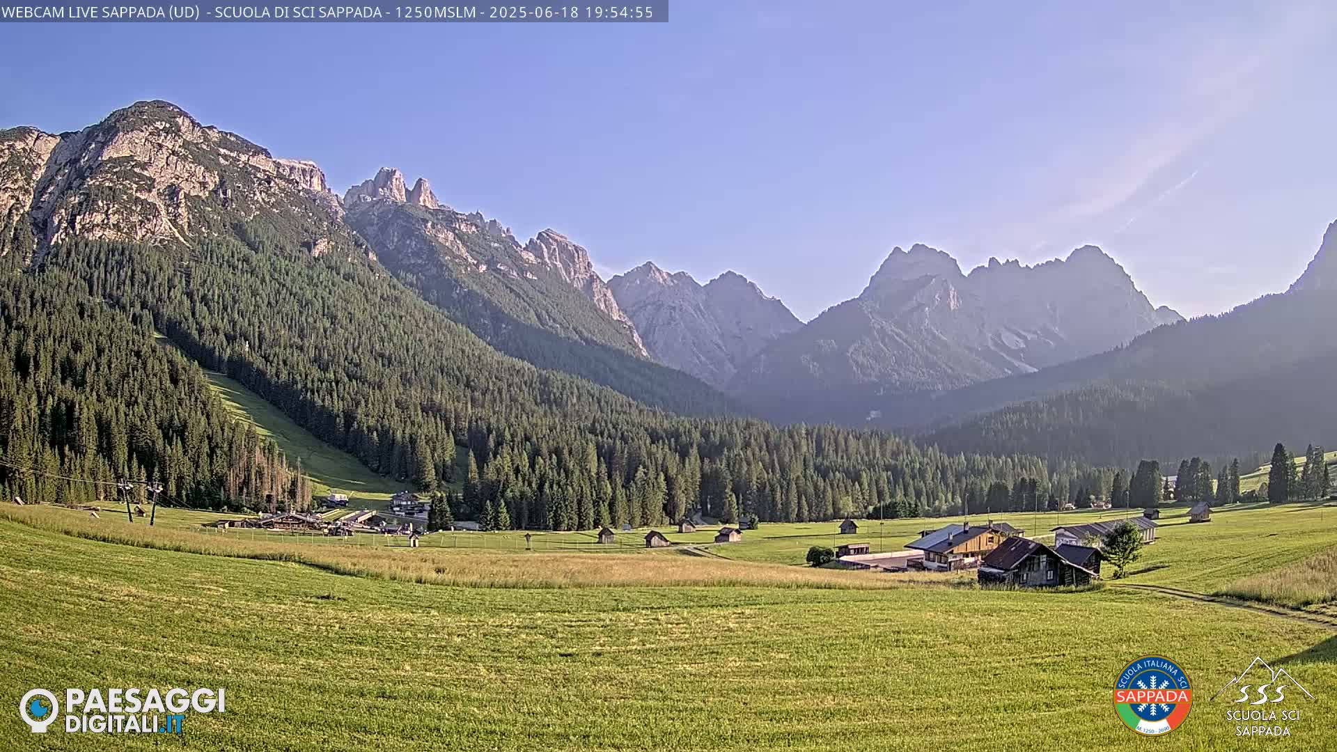 A valley with green fields, a few buildings, and a dark evergreen forest is nestled beneath a range of light-grey, rocky mountains under a clear, light-blue sky.