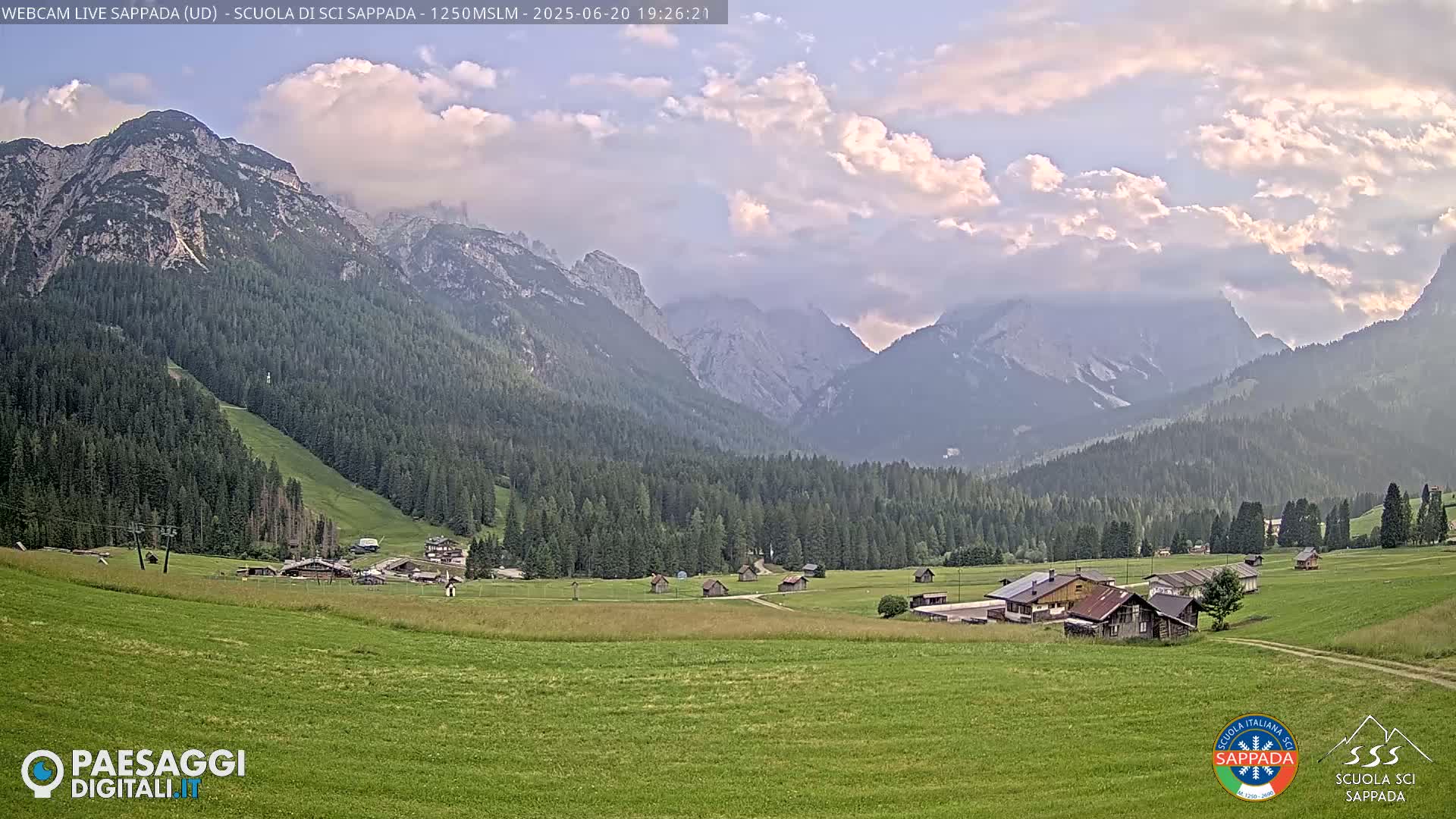 A valley with green fields and a small village is nestled between towering mountains under a partly cloudy sky.