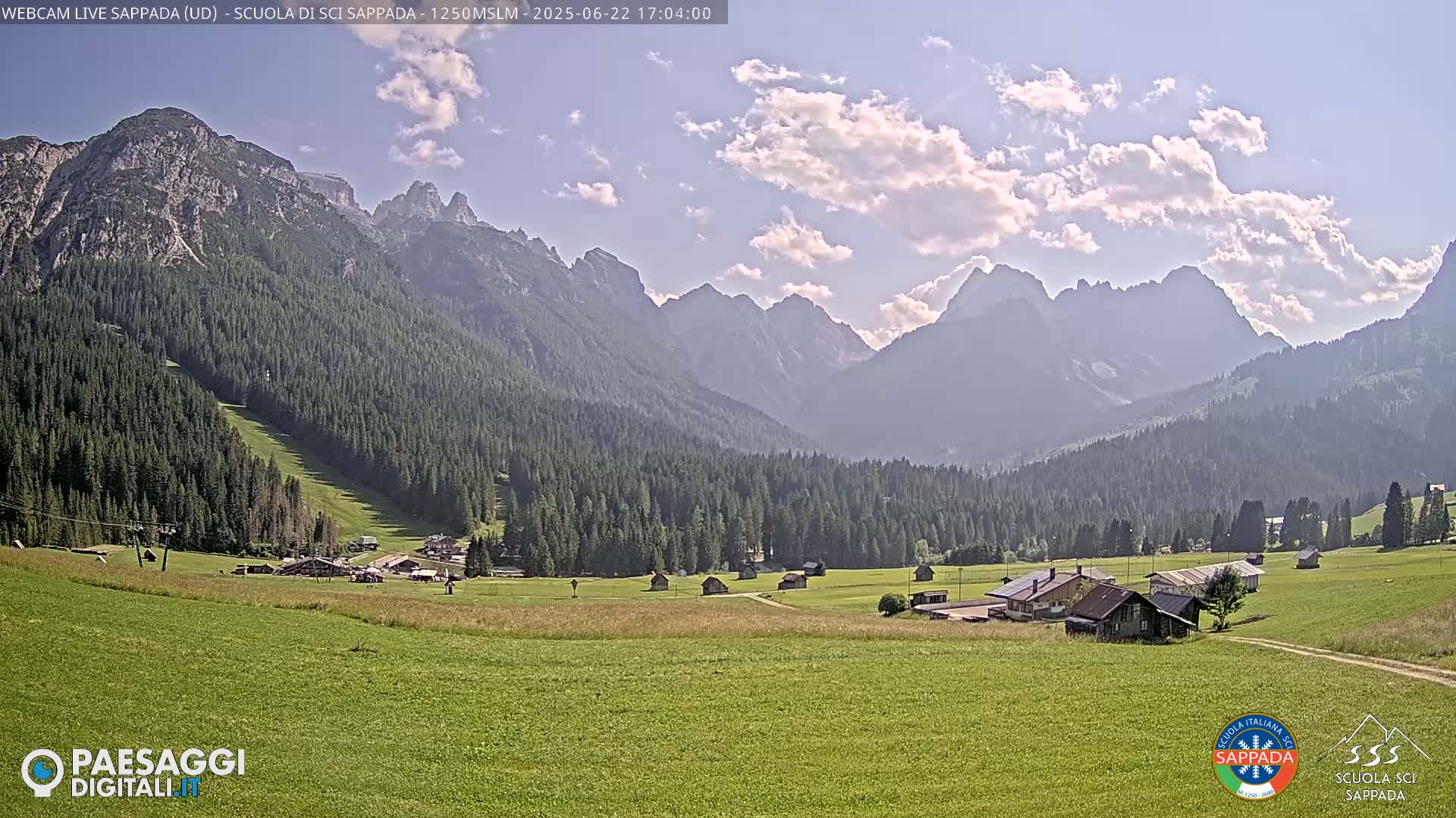 A sunny day reveals a valley of green fields and dark evergreen forests nestled between towering, hazy mountains under a partly cloudy sky.