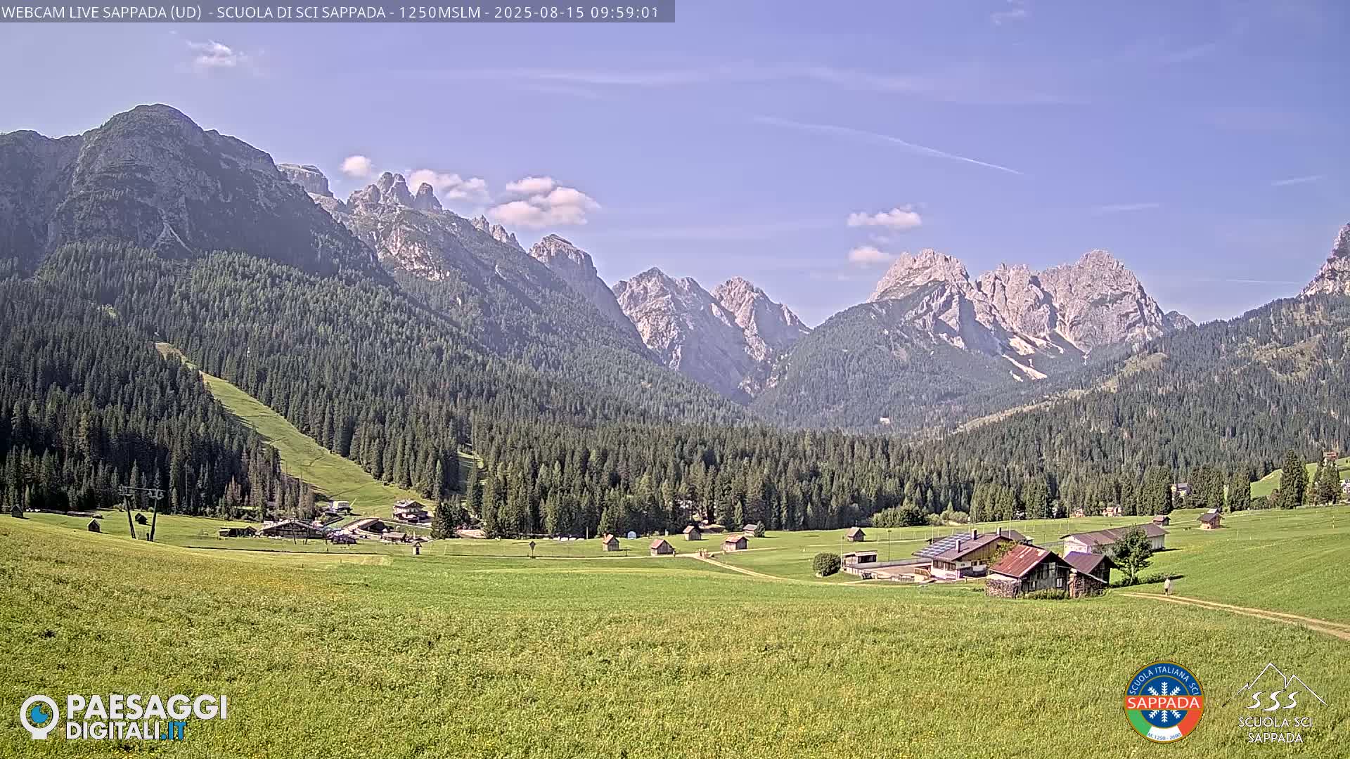 A sunny day reveals a valley of green fields and dark evergreen forests nestled among towering, rocky mountains.