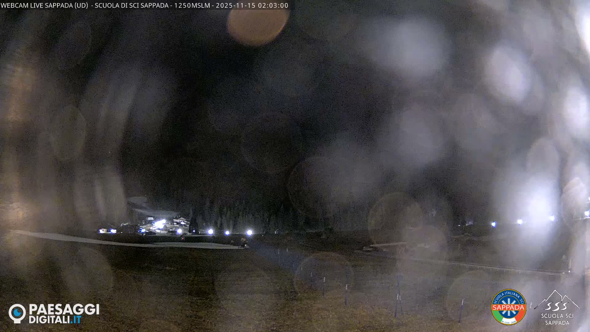 A heavily blurred night-time image shows brightly lit snowmaking machinery on a ski slope, with low visibility caused by dense fog or heavy snowfall and moisture on the camera lens.