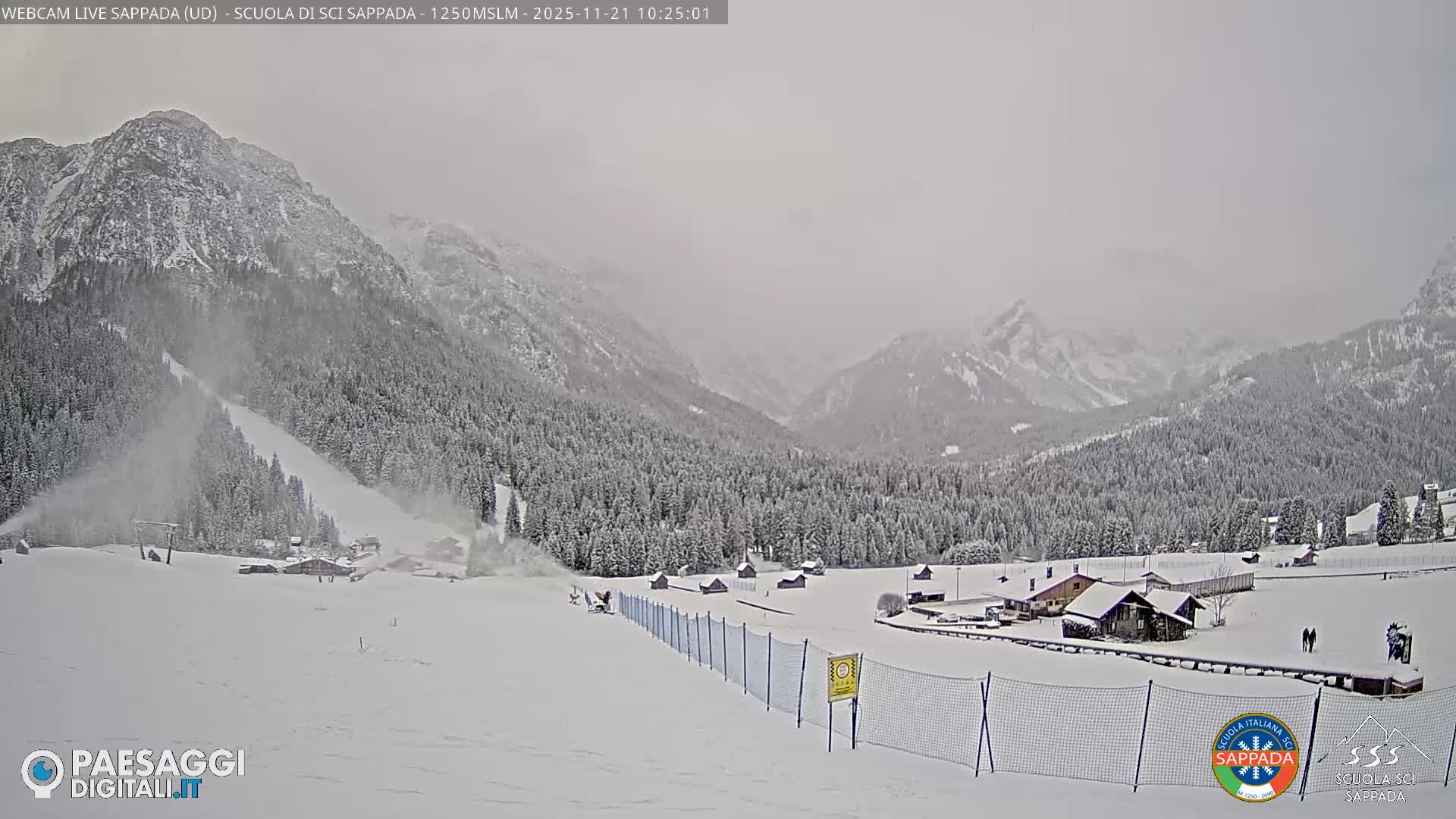 A wide shot reveals a snow-covered mountain valley with a ski slope being prepared by a snow cannon, surrounded by dense evergreen forests and scattered buildings under a gray, overcast sky.