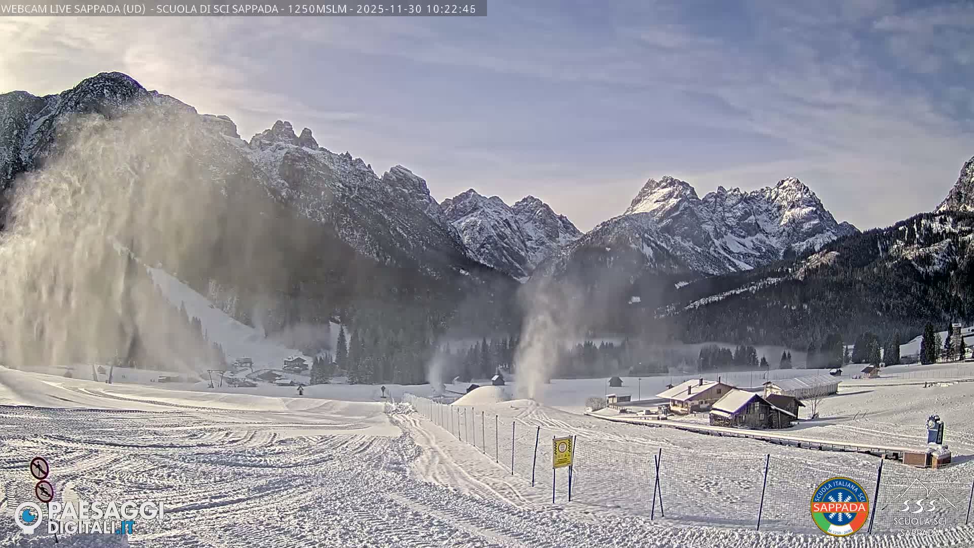 Snow cannons actively spray artificial snow across wide, groomed ski slopes in a snowy mountain valley with pine forests and scattered buildings, all beneath a partly cloudy winter sky and surrounded by majestic snow-capped peaks.