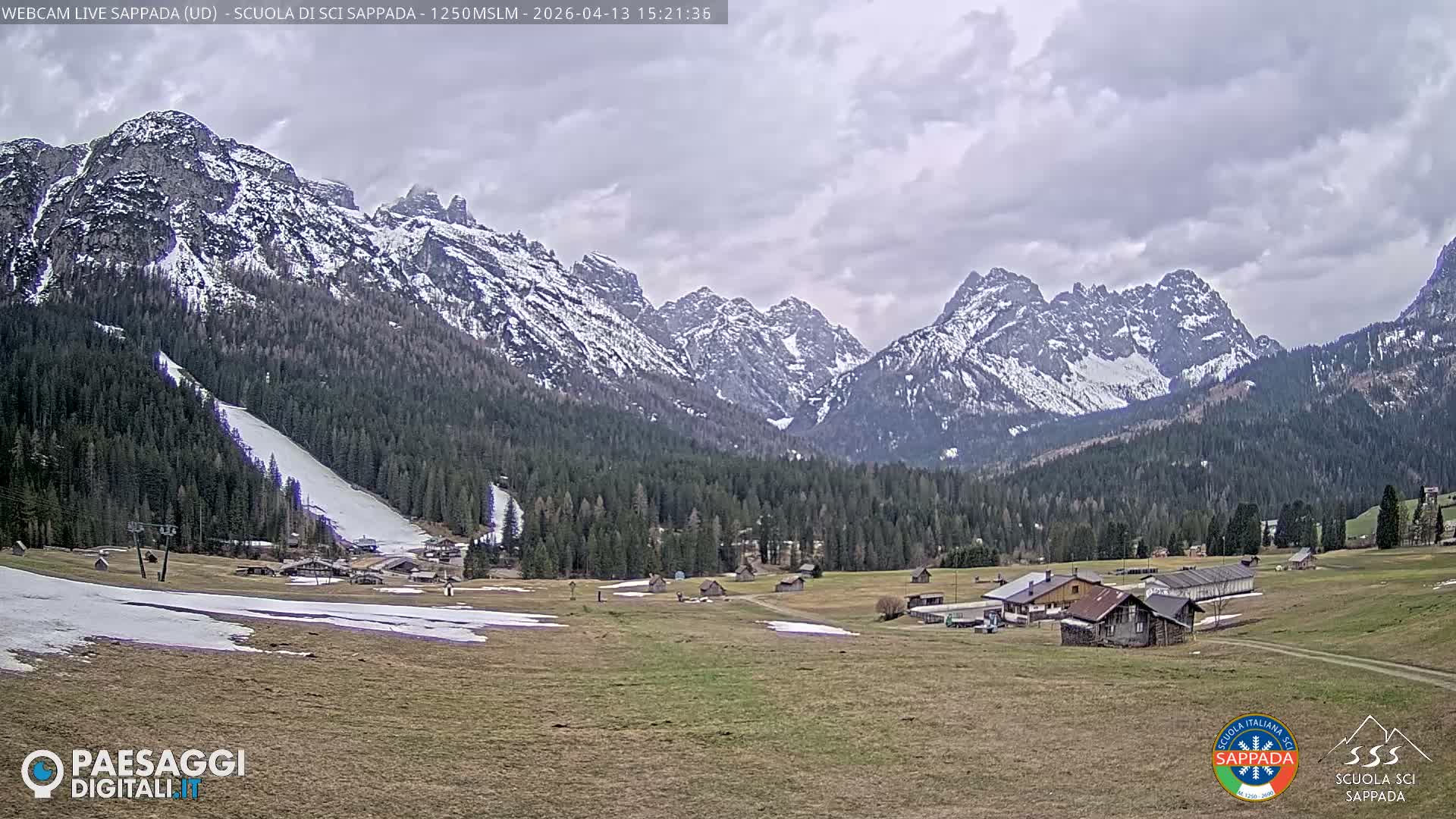 A wide panoramic view captures a snow-covered mountain range towering over dense pine forests and a valley featuring small buildings, a ski slope, and a fenced-off snowy field, all under an overcast sky.