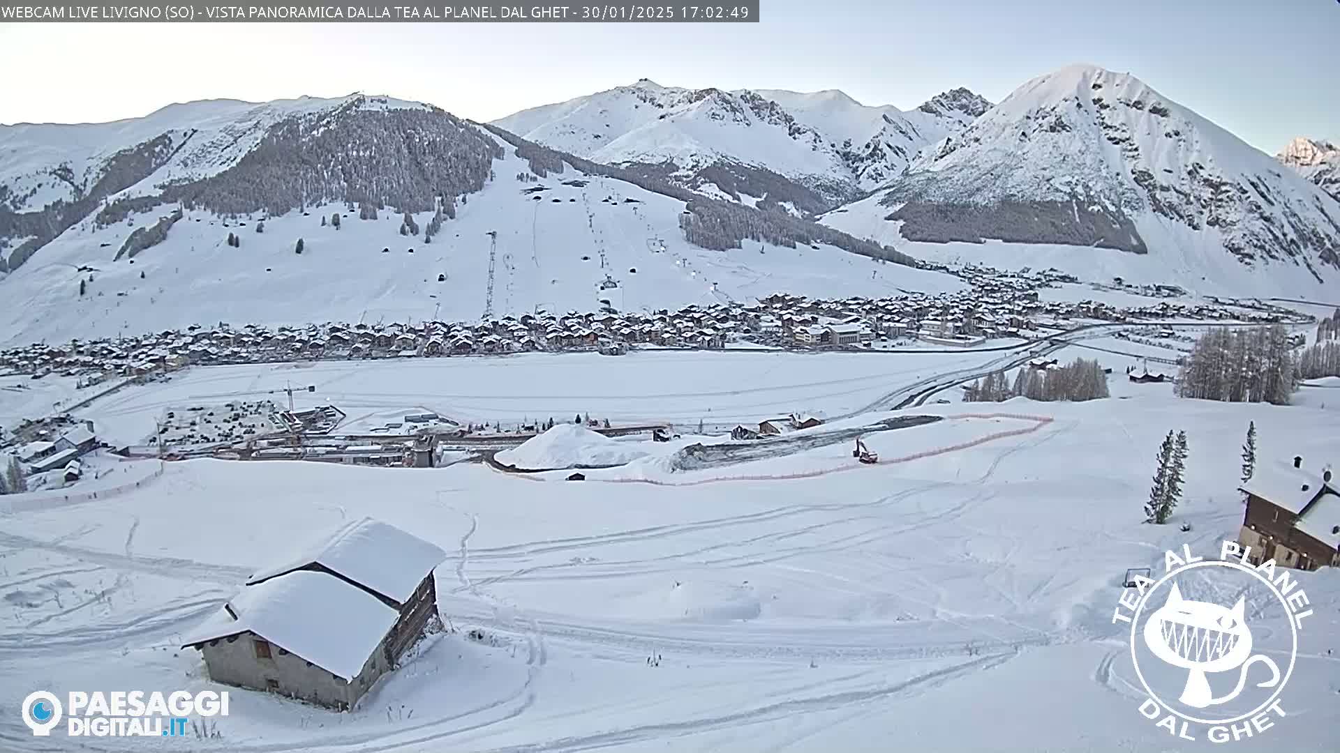 A snow-covered mountain village is nestled in a valley beneath snow-capped mountains on a clear day.