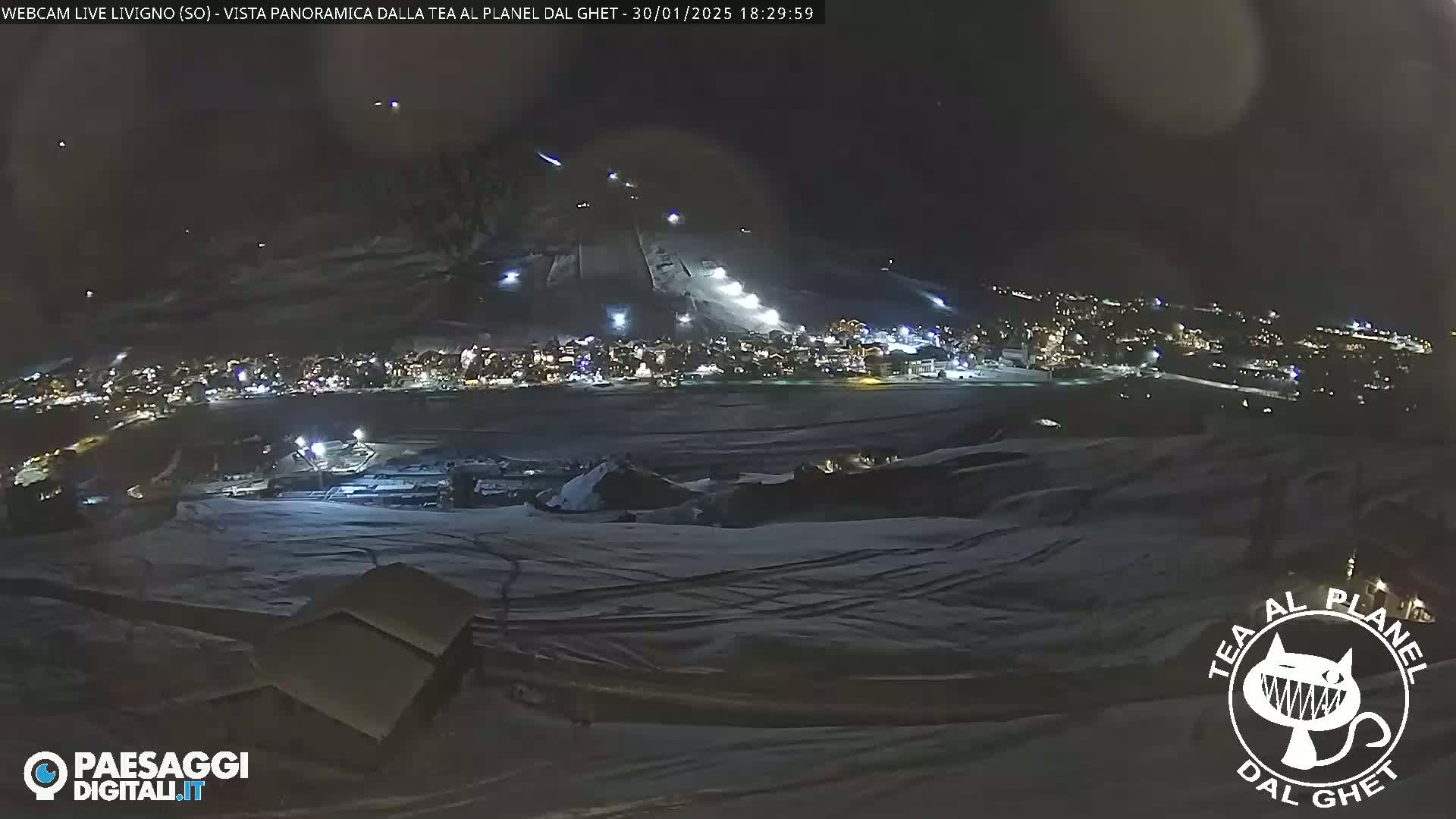 A nighttime, snow-covered landscape shows a brightly lit town nestled below a ski slope with several skiers visible.