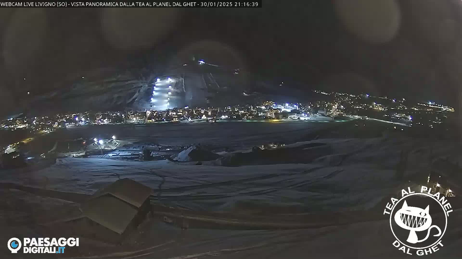 A nighttime, snow-covered landscape with a brightly lit ski slope and a town visible in the distance under a dark sky.