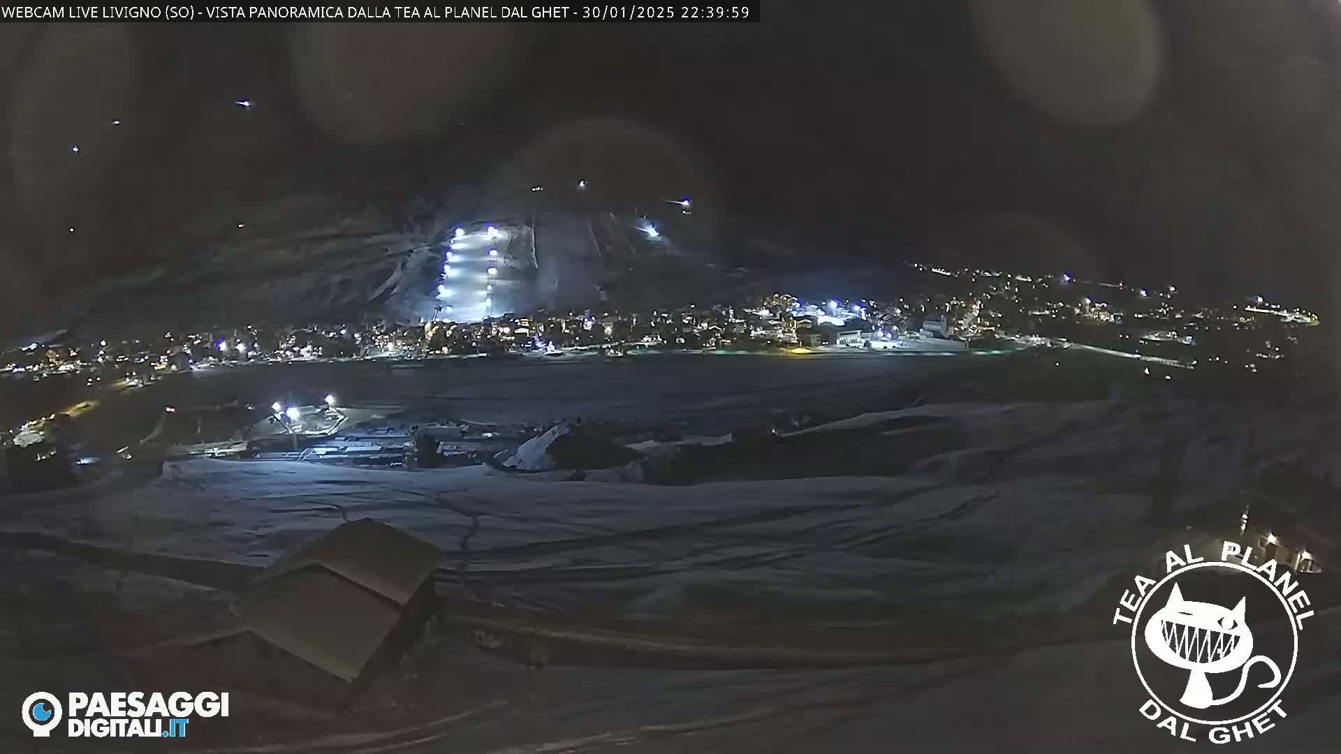 A nighttime, snow-covered landscape shows a town illuminated below a ski slope with lights on the runs.