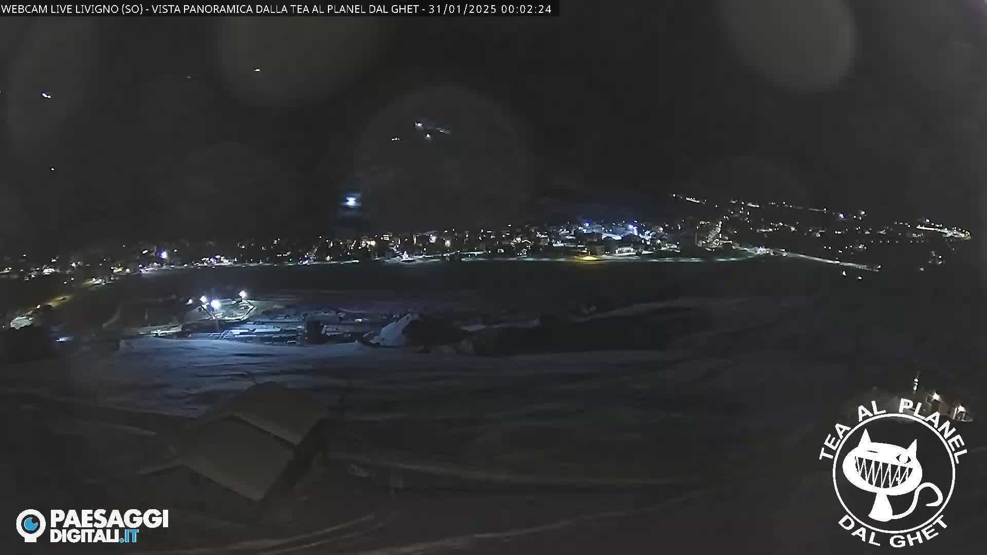A nighttime panoramic view of a snow-covered landscape, showing a town with lights in the distance under a dark, possibly slightly hazy sky.