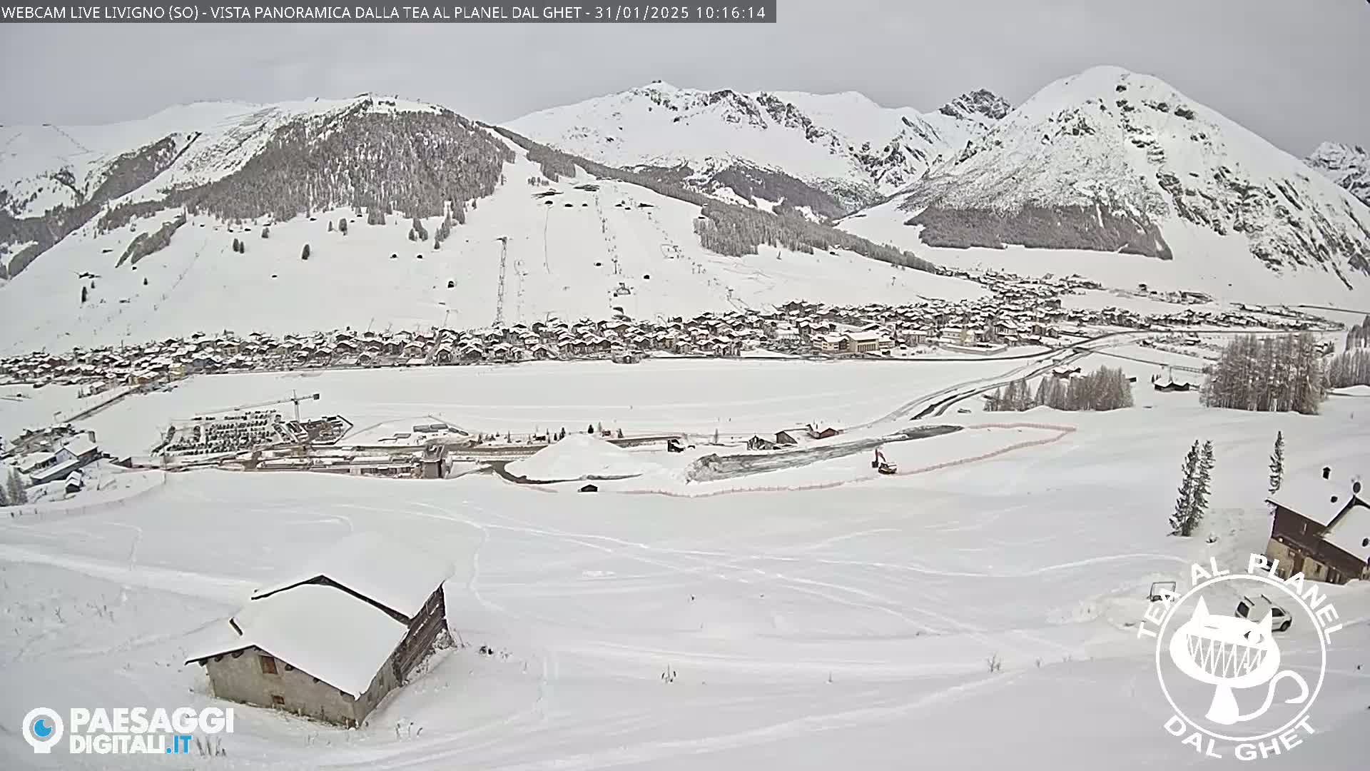 A snow-covered mountain village is nestled in a valley surrounded by snow-capped mountains under an overcast sky.