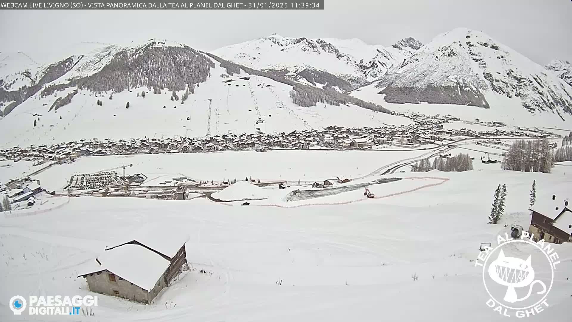 A snow-covered mountain village is seen from a high vantage point on a cloudy day, with a small building in the foreground and snow-covered slopes and mountains in the background.