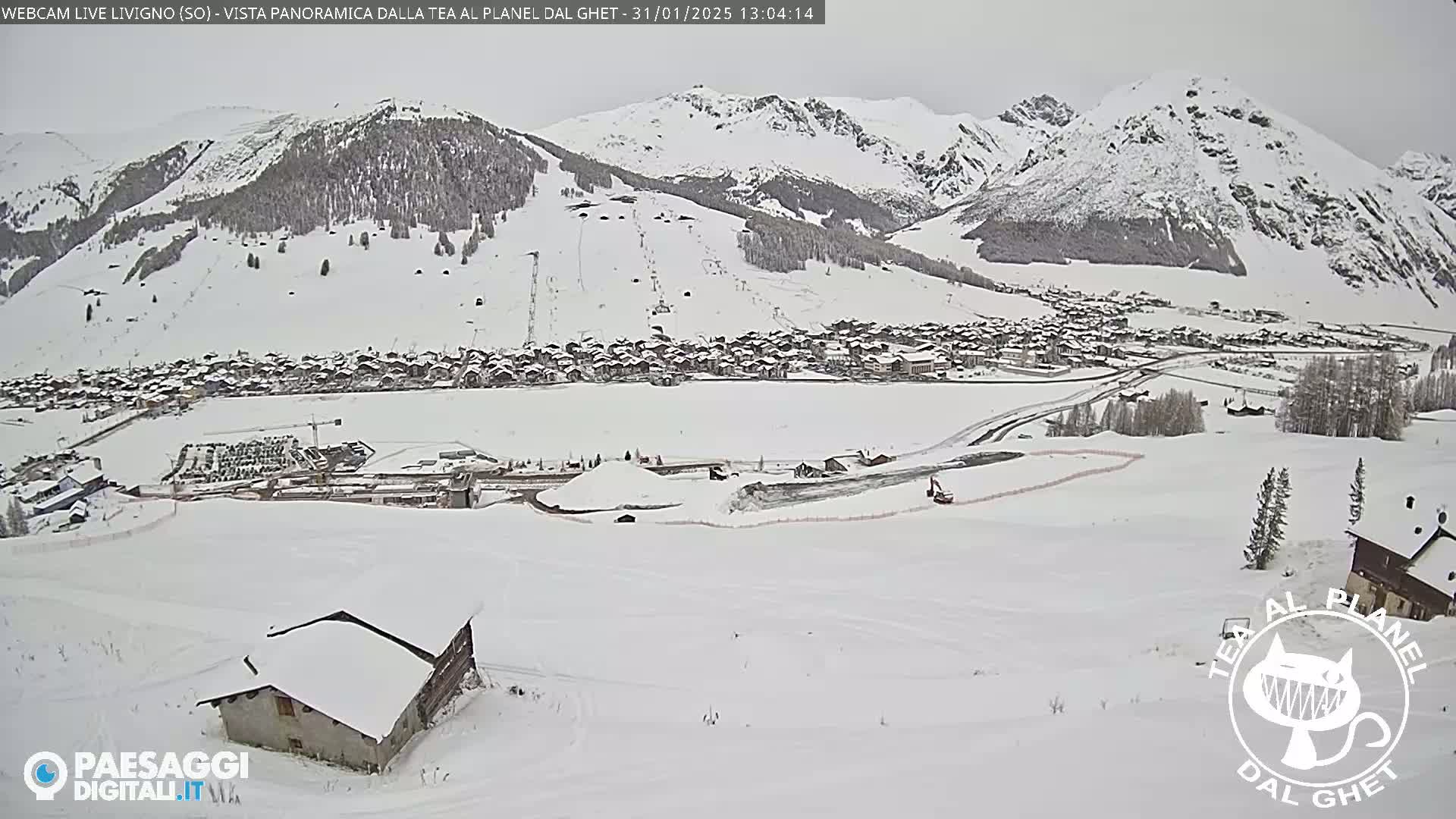 A snow-covered village nestled in a valley between snow-capped mountains, viewed from a higher elevation on a cloudy day.