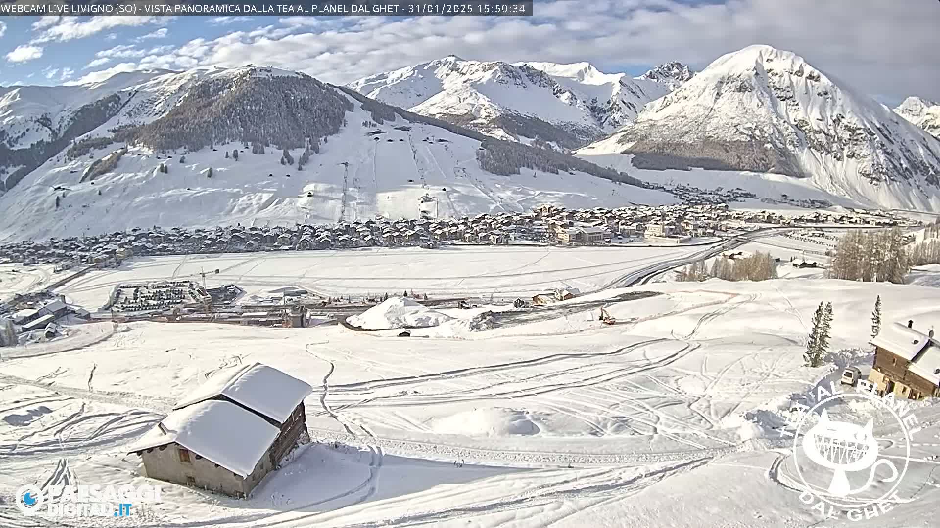 A snow-covered mountain village is viewed from above on a partly sunny day, with snow-covered mountains in the background and tire tracks visible in the foreground.