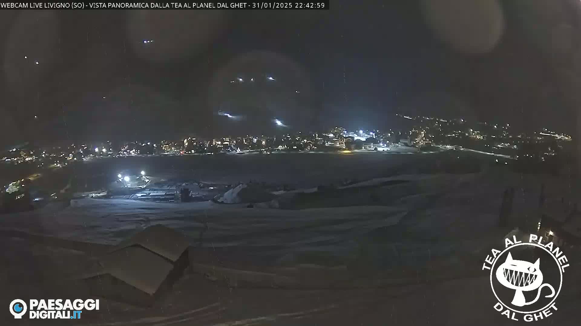 A nighttime, snow-covered landscape shows a town illuminated in the distance under a dark sky with streaks of light possibly from fireworks or aircraft contrails.