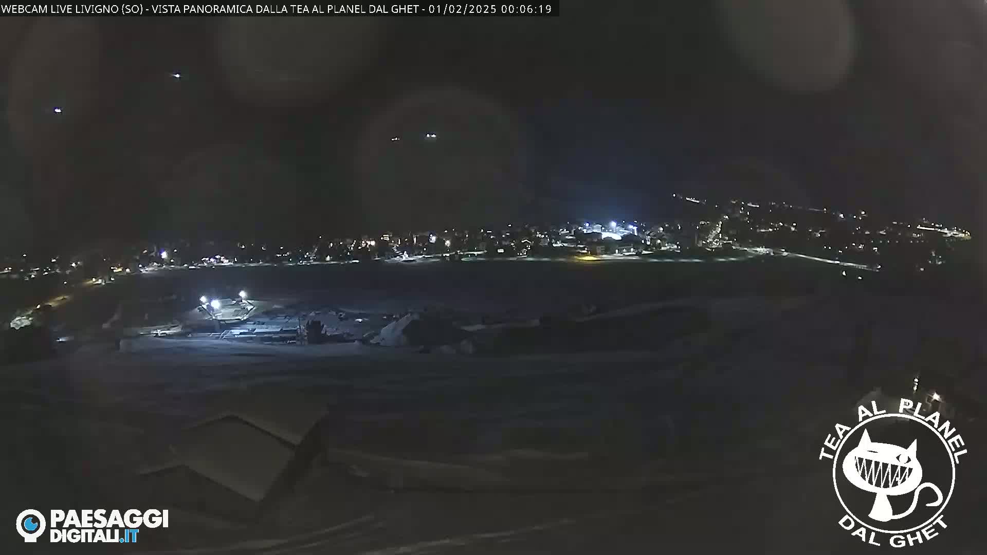 A nighttime, panoramic view of a snow-covered landscape shows a distant town illuminated with lights under a clear, dark sky.
