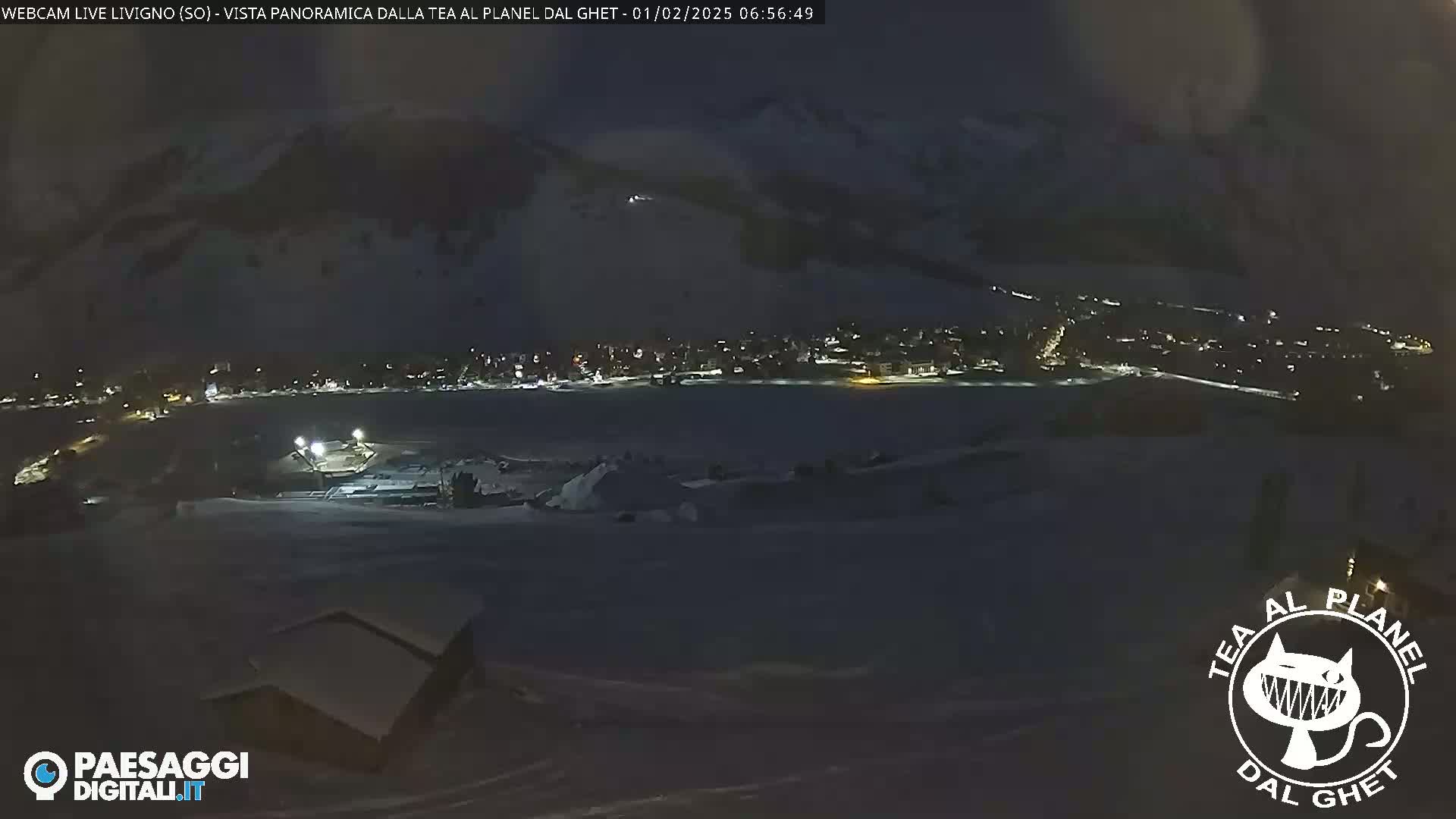 A nighttime, panoramic view of a snow-covered town nestled in a valley, illuminated by scattered lights, with mountains in the background under a dark sky.