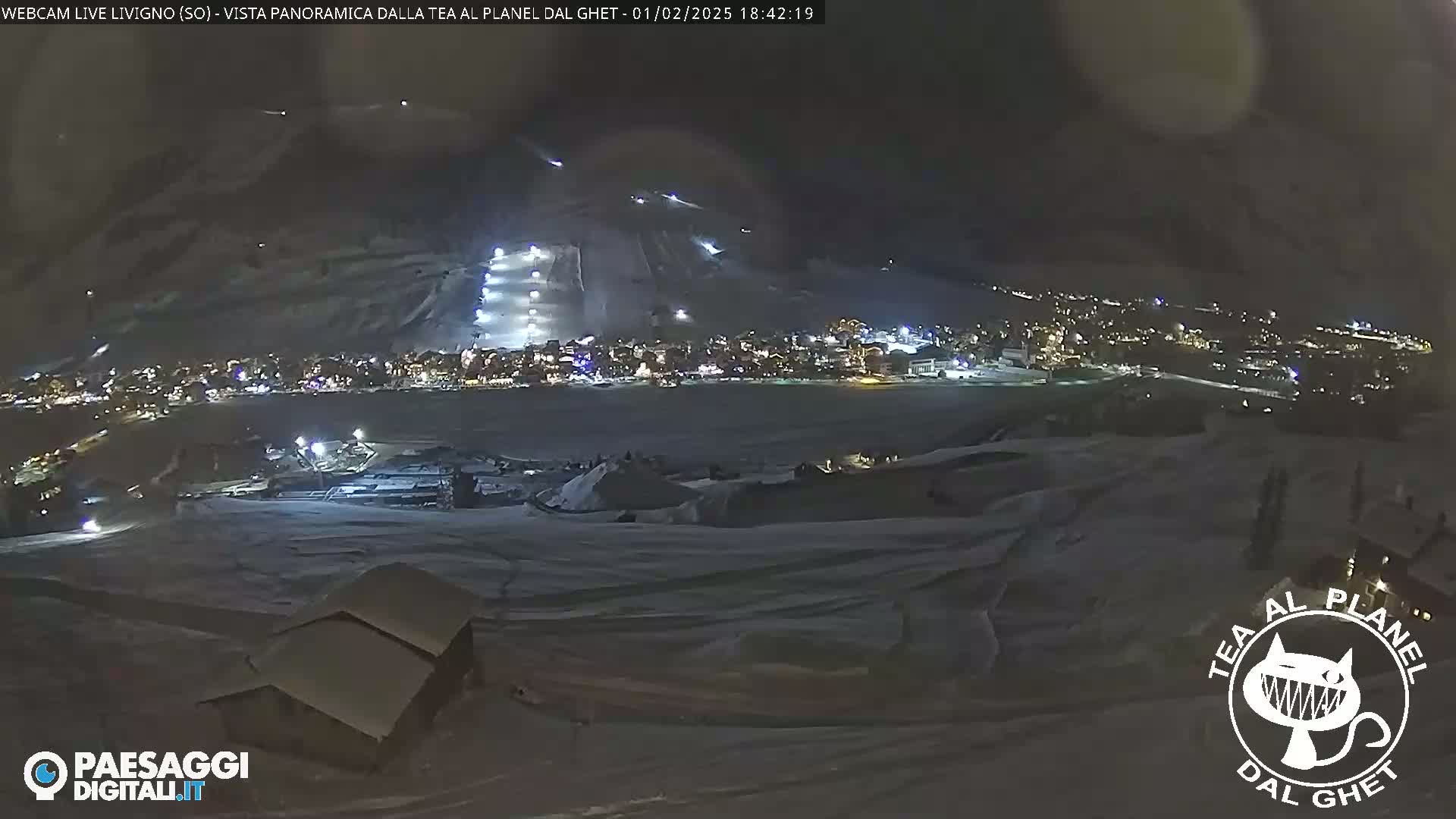 A nighttime, snow-covered landscape shows a town illuminated below a brightly lit ski slope.