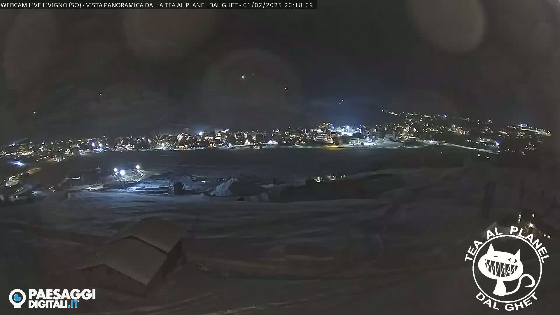 A nighttime, panoramic view of a snow-covered landscape shows a town with lights in the distance under a dark, cloudy sky.