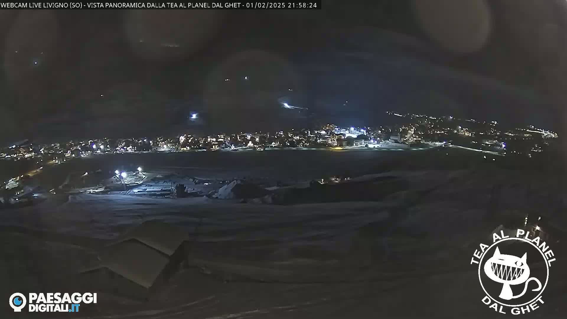 A nighttime, snowy landscape shows a town illuminated below, with streaks of light possibly from aircraft in the dark sky.