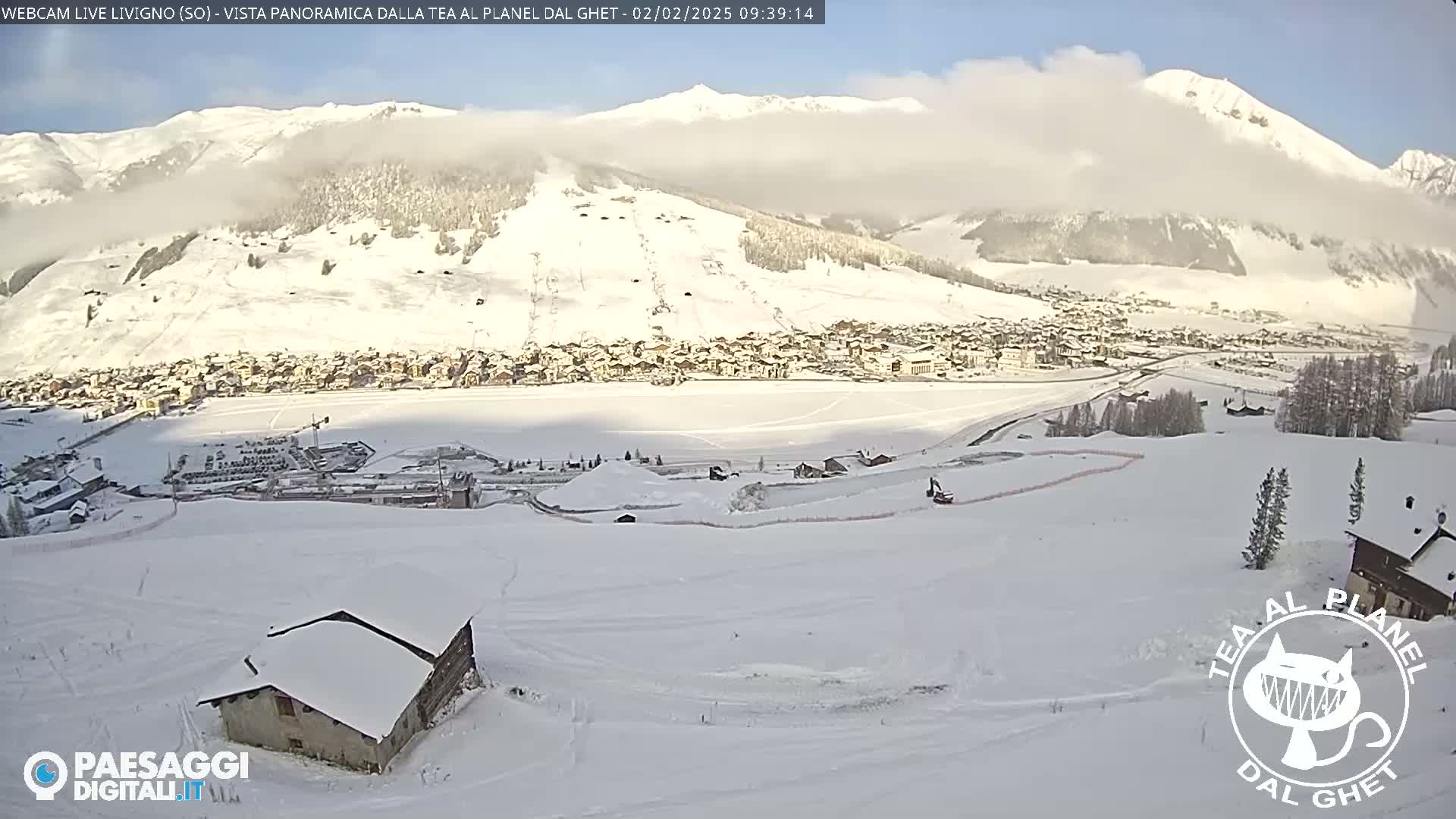 A snow-covered village nestled in a valley surrounded by snow-capped mountains under a mostly clear, sunny sky.