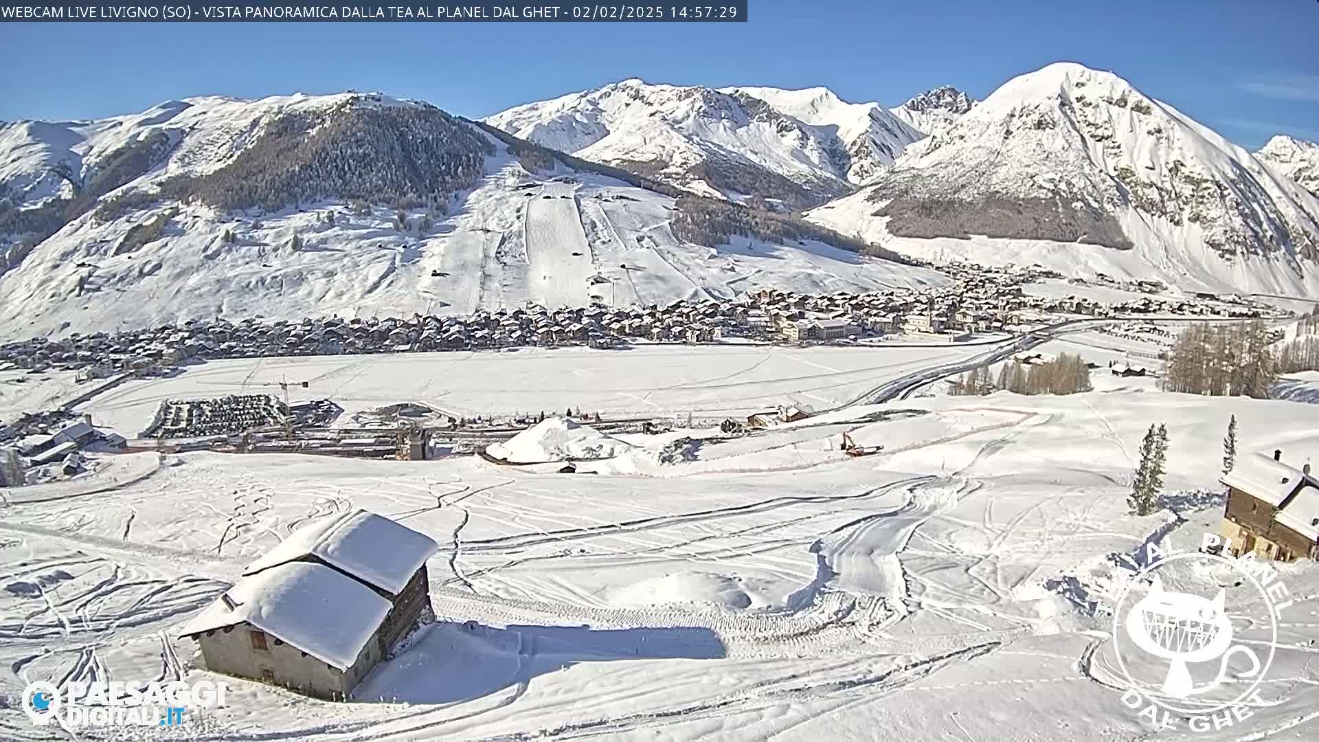 A snow-covered mountain village is viewed from above on a sunny day, with ski slopes and a small house visible in the foreground.
