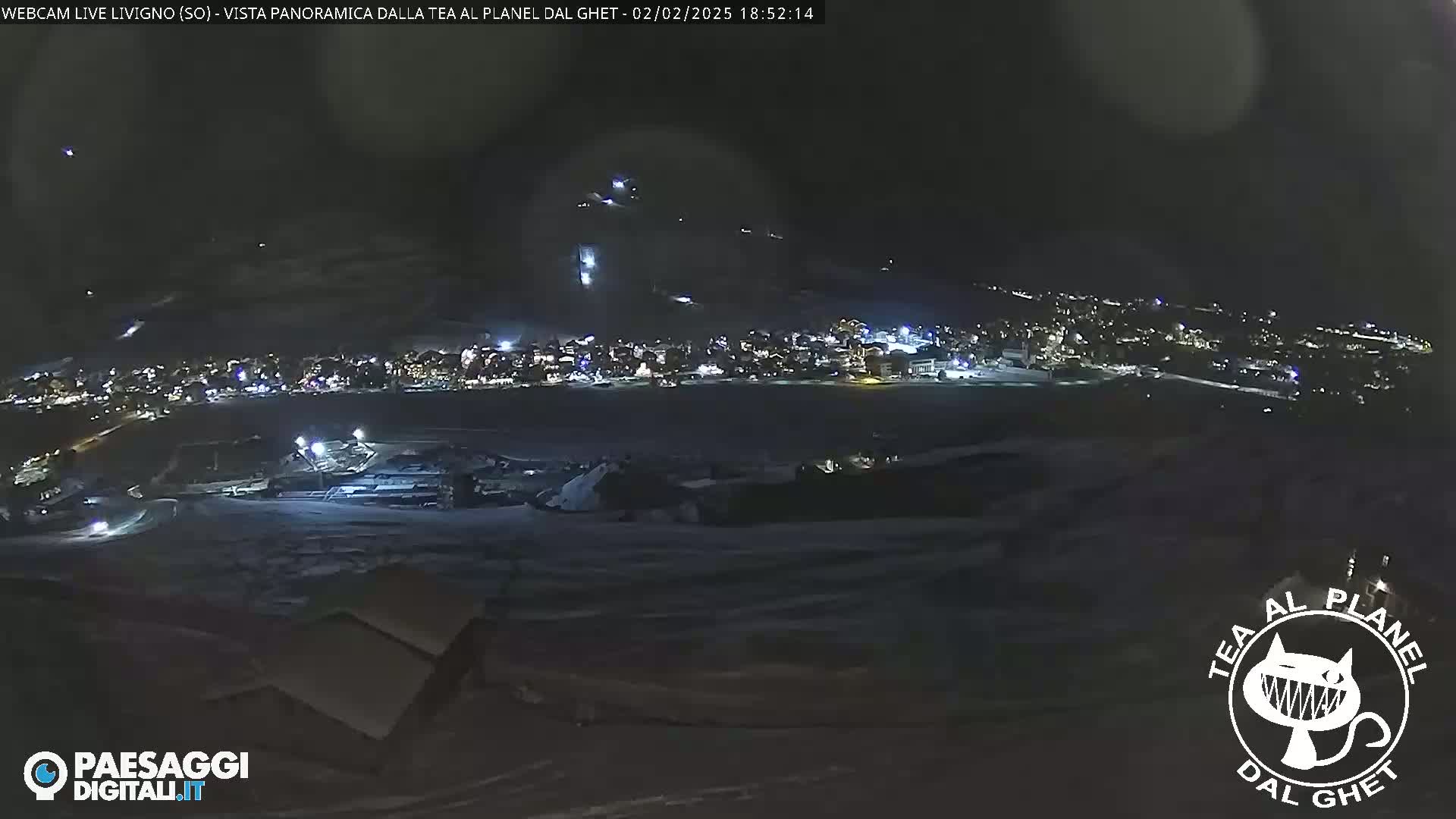 A nighttime long shot of a snow-covered town with lights illuminated, situated below a dark, snow-covered hillside with faint lights from what appears to be a ski lift.