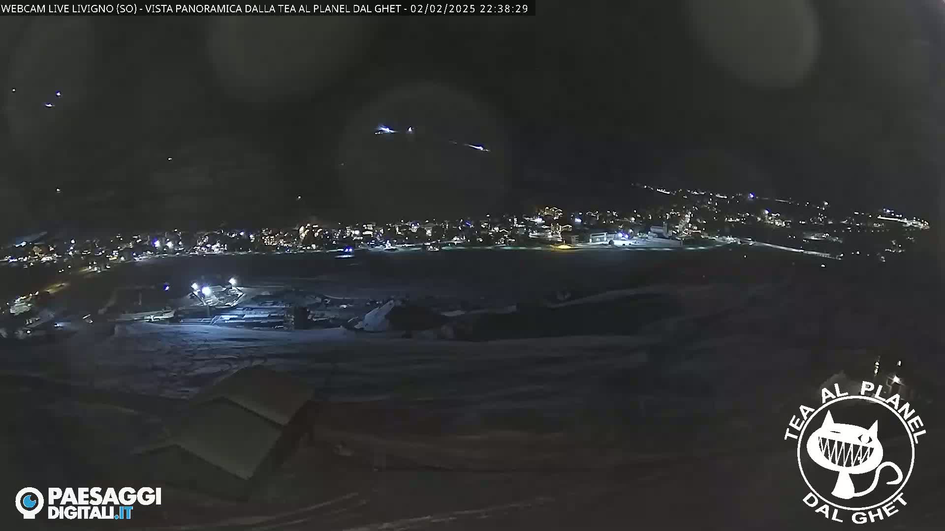 A nighttime, panoramic view of a snow-covered town with lights illuminated, seen from a high vantage point under a clear, dark sky.