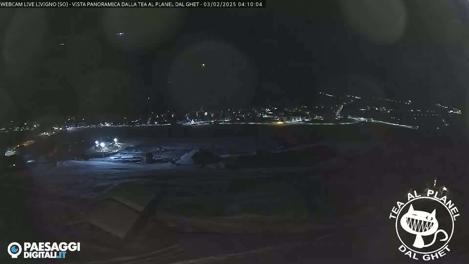 A nighttime, panoramic view of a snow-covered landscape with a town's lights visible in the distance under a dark, clear sky.